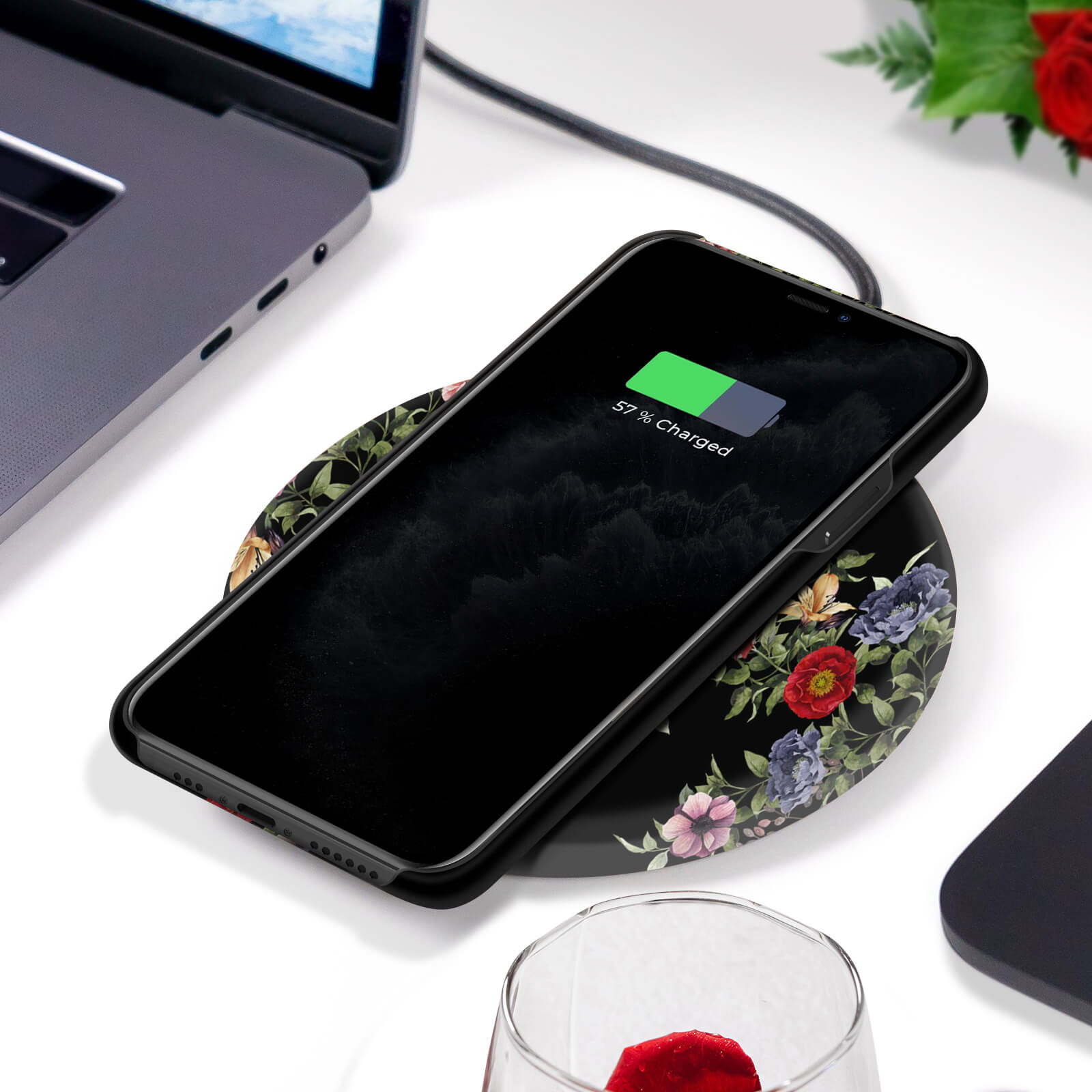 Telefon auf floralem Ladegerät. Lädt mit 57%. Schwarze Hülle mit Blumendesign. Weiße Oberfläche.