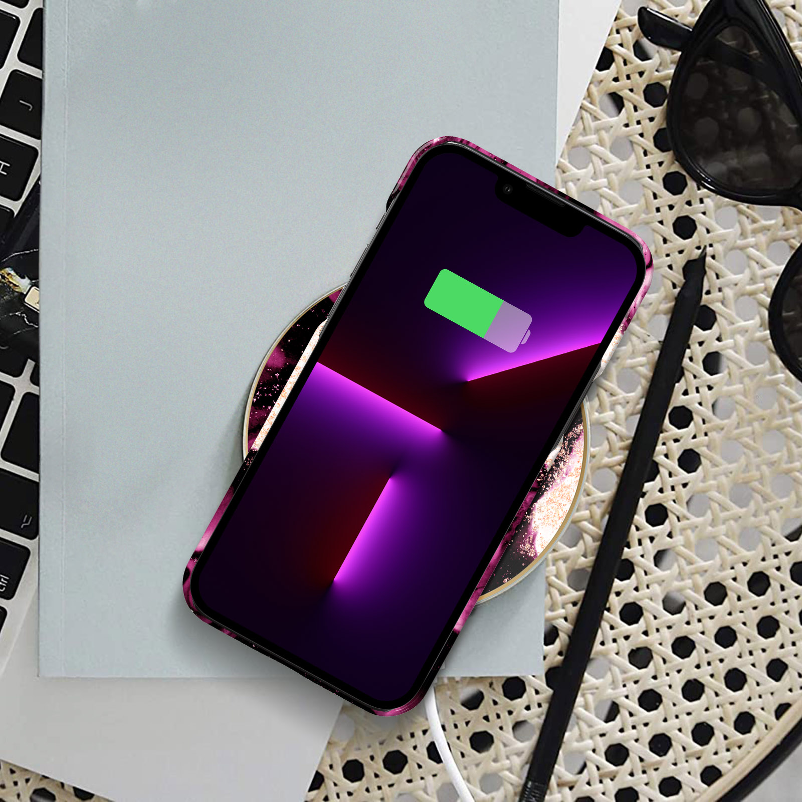 Kablosuz bir şarj cihazında şarj olan bir telefon. Telefonun pembe bir kılıfı ve yeşil bir pil simgesi var. Güneş gözlüğü.
