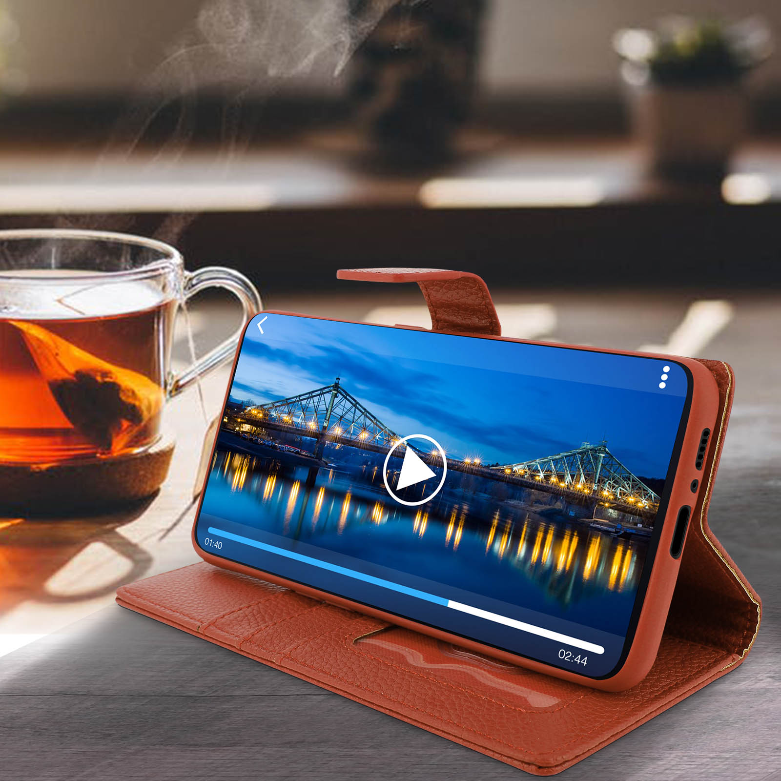 Smartphone in custodia marrone, riproduce un video con scena del ponte, tazza di tè accanto.