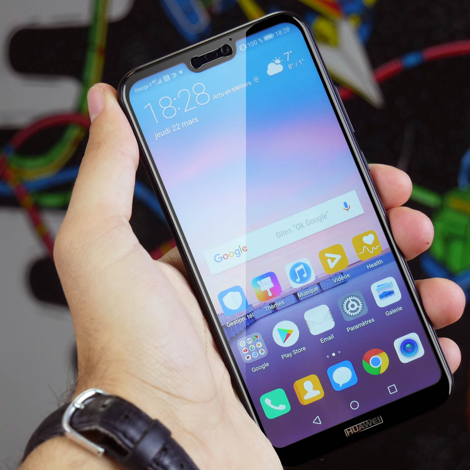 Une main tenant un smartphone avec des icônes d'applications. La date et l'heure sont affichées. Le fond est flou.
