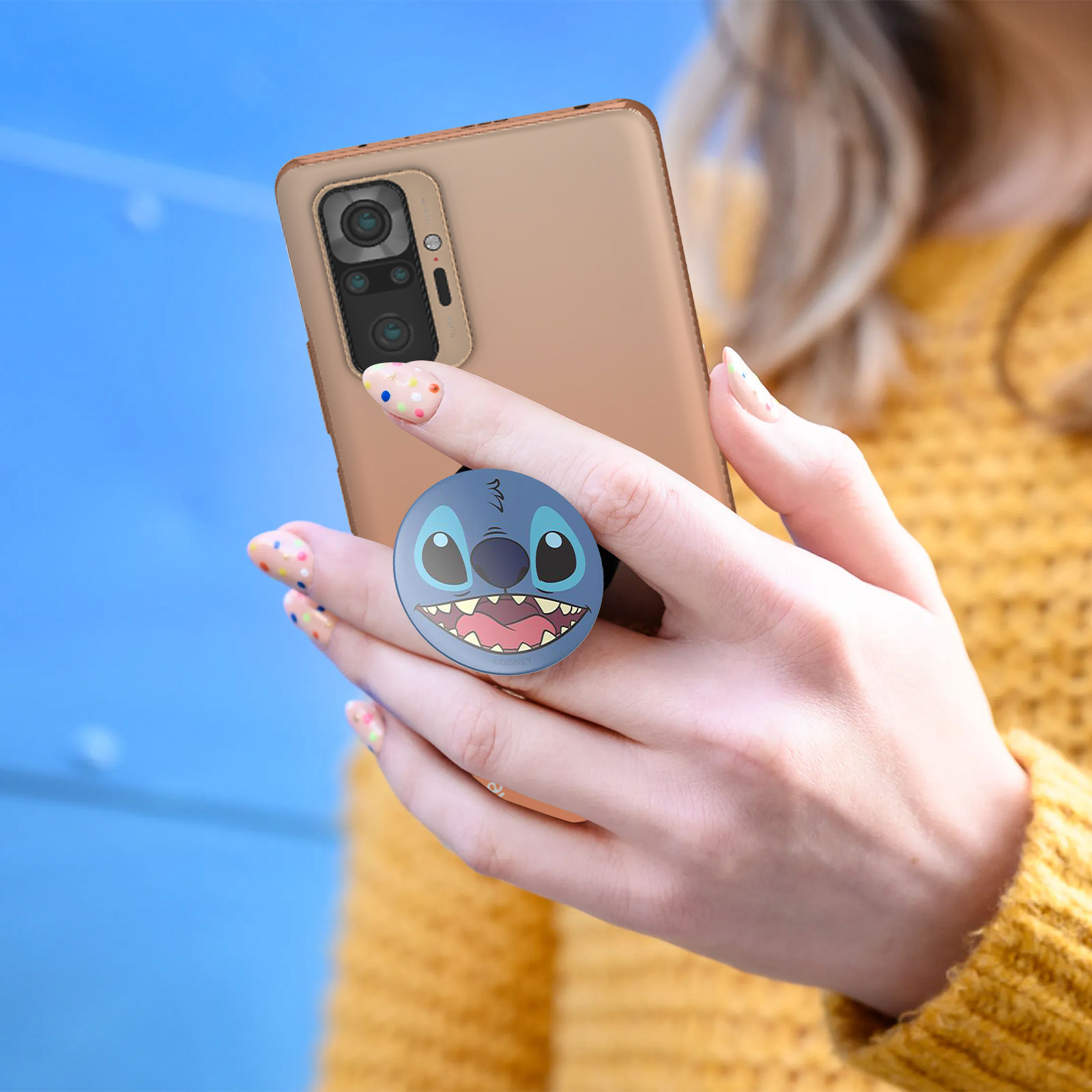 Eine Frau hält ein Telefon mit einem Stitch Pop-Socket, mit gelbem Pullover.