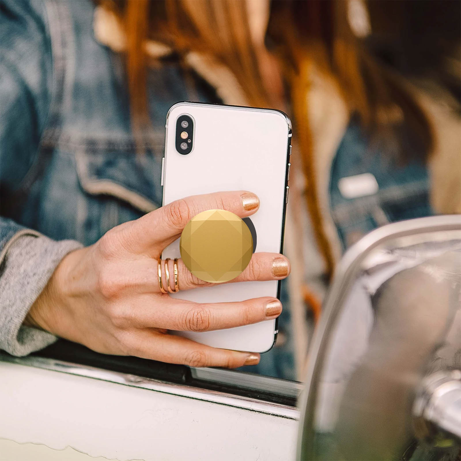 Eine Person hält ein weißes Telefon mit einem goldenen PopSocket. Die Person trägt Ringe und hat goldenen Nagellack.