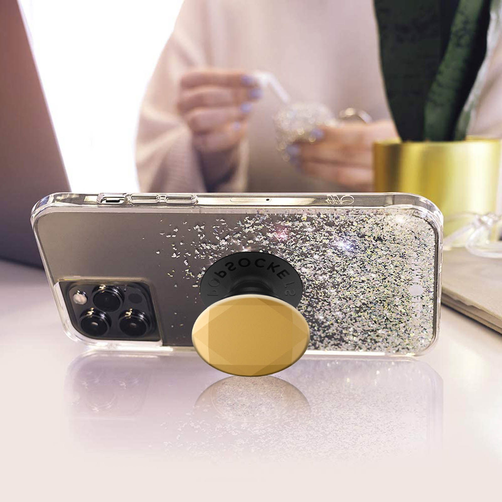 Ein Telefon mit PopSocket auf einem Tisch. Glitzer ist auf dem Telefon. Hände im Hintergrund.