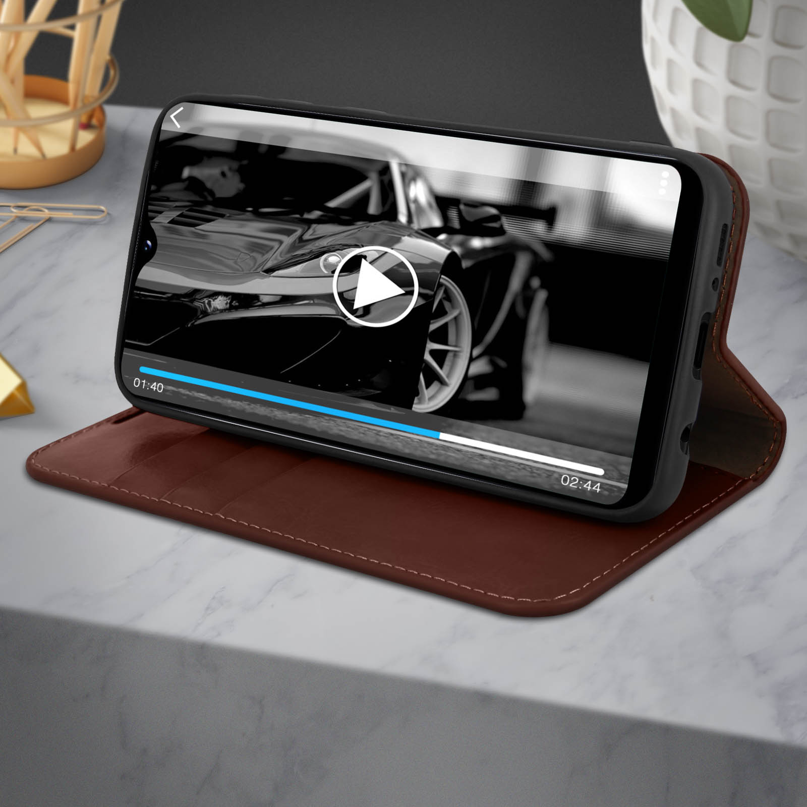 Telefon zeigt ein Autovideo. Es befindet sich in einem braunen Etui auf einer Marmoroberfläche. Eine goldene Büroklammer ist sichtbar.
