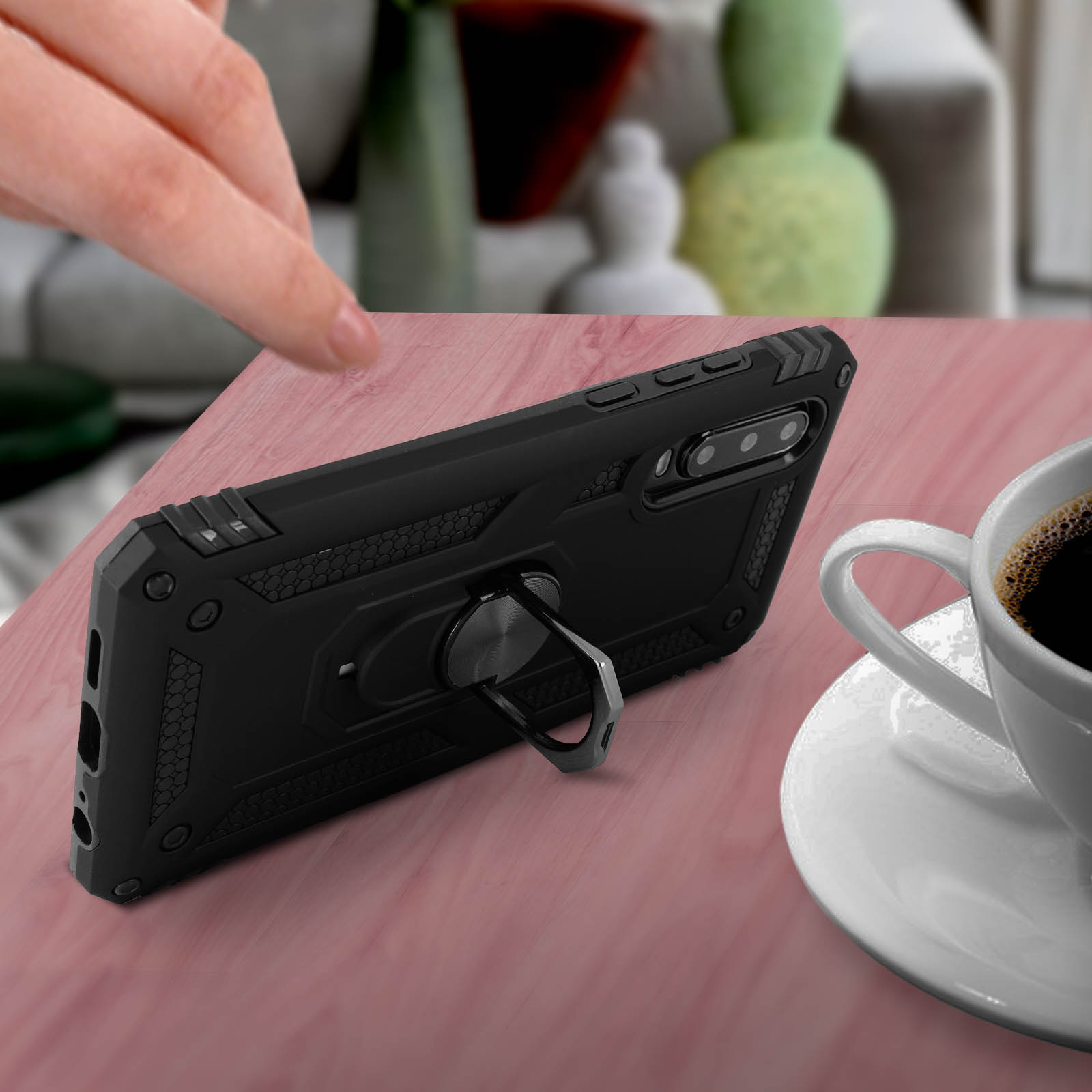 Un téléphone noir avec un support de bague sur une surface rose, à côté d'une tasse de café.