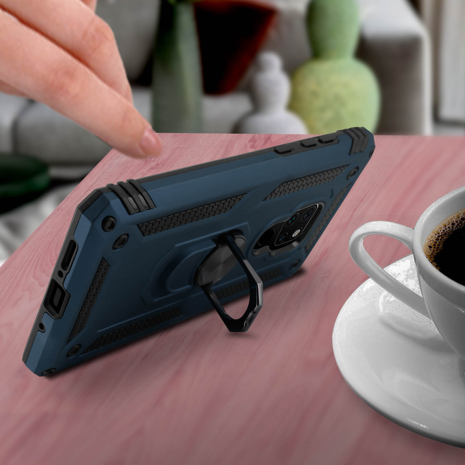 Una custodia blu per telefono con supporto ad anello è su un tavolo rosa con una tazza di caffè.
