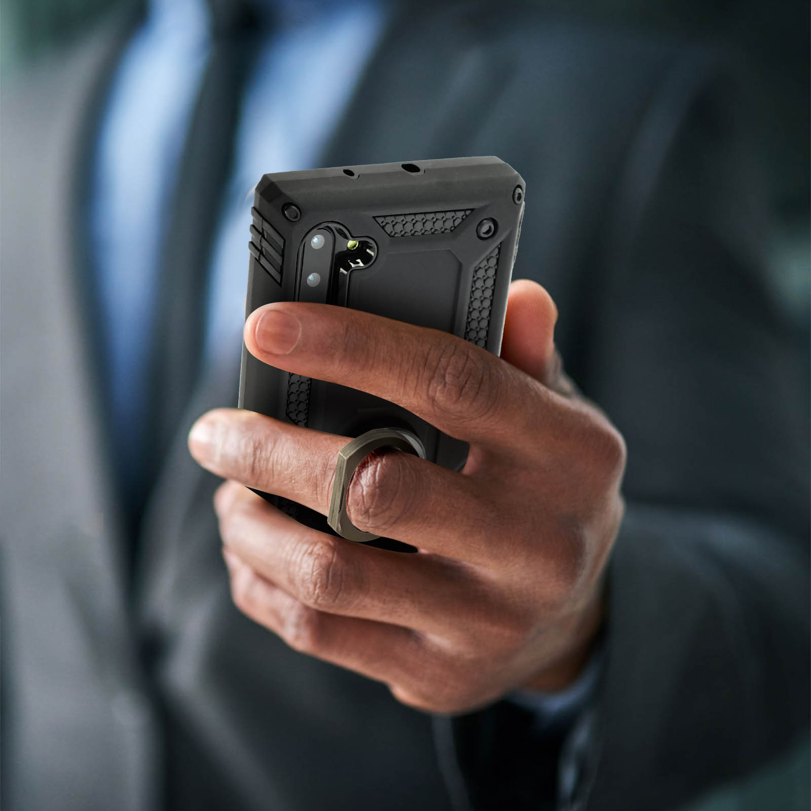 Una mano tiene un telefono nero. Il telefono ha una custodia nera con un supporto ad anello. Uomo in giacca e cravatta sullo sfondo.