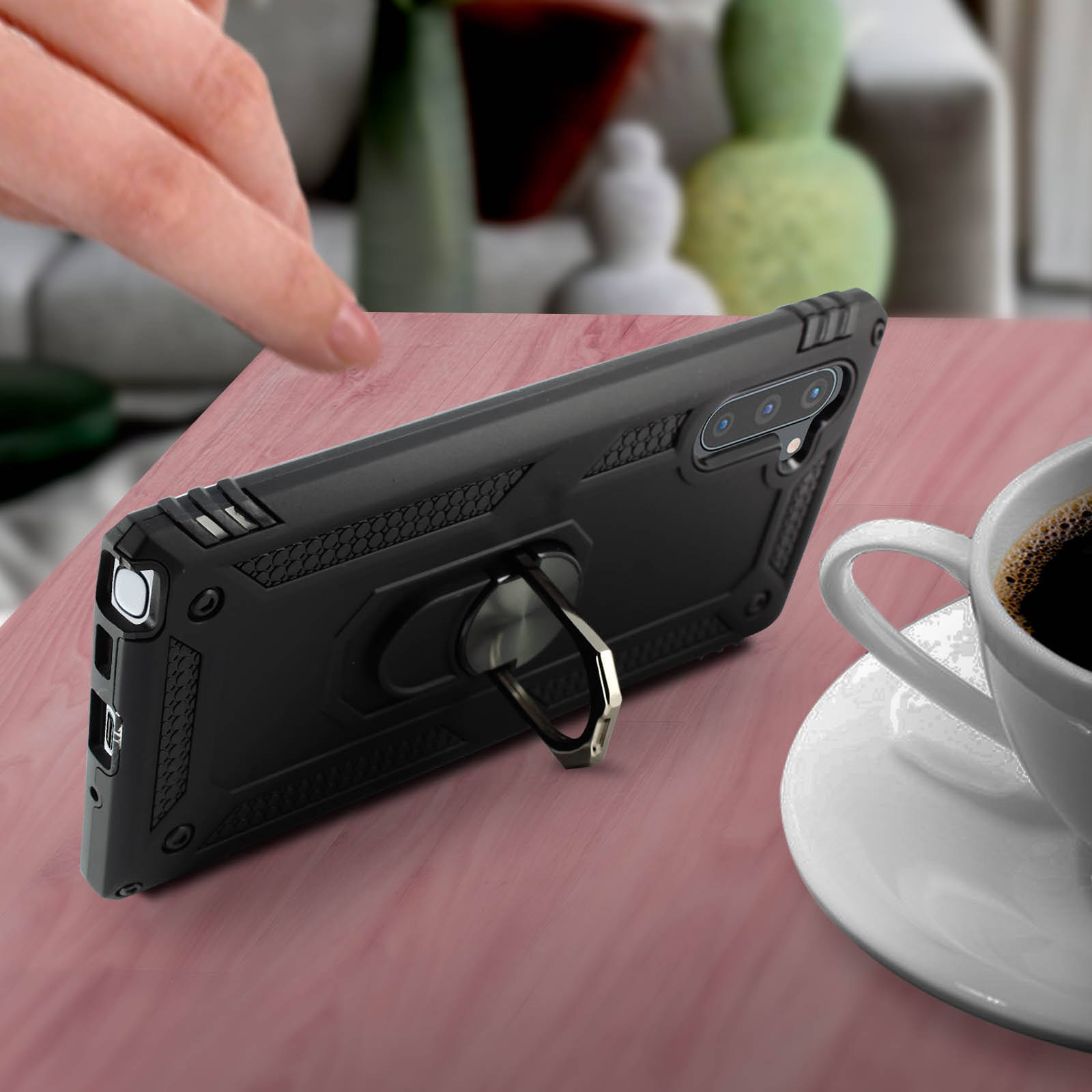 Una custodia nera per telefono con un supporto ad anello su un tavolo di legno accanto a una tazza di caffè. Una mano è visibile.