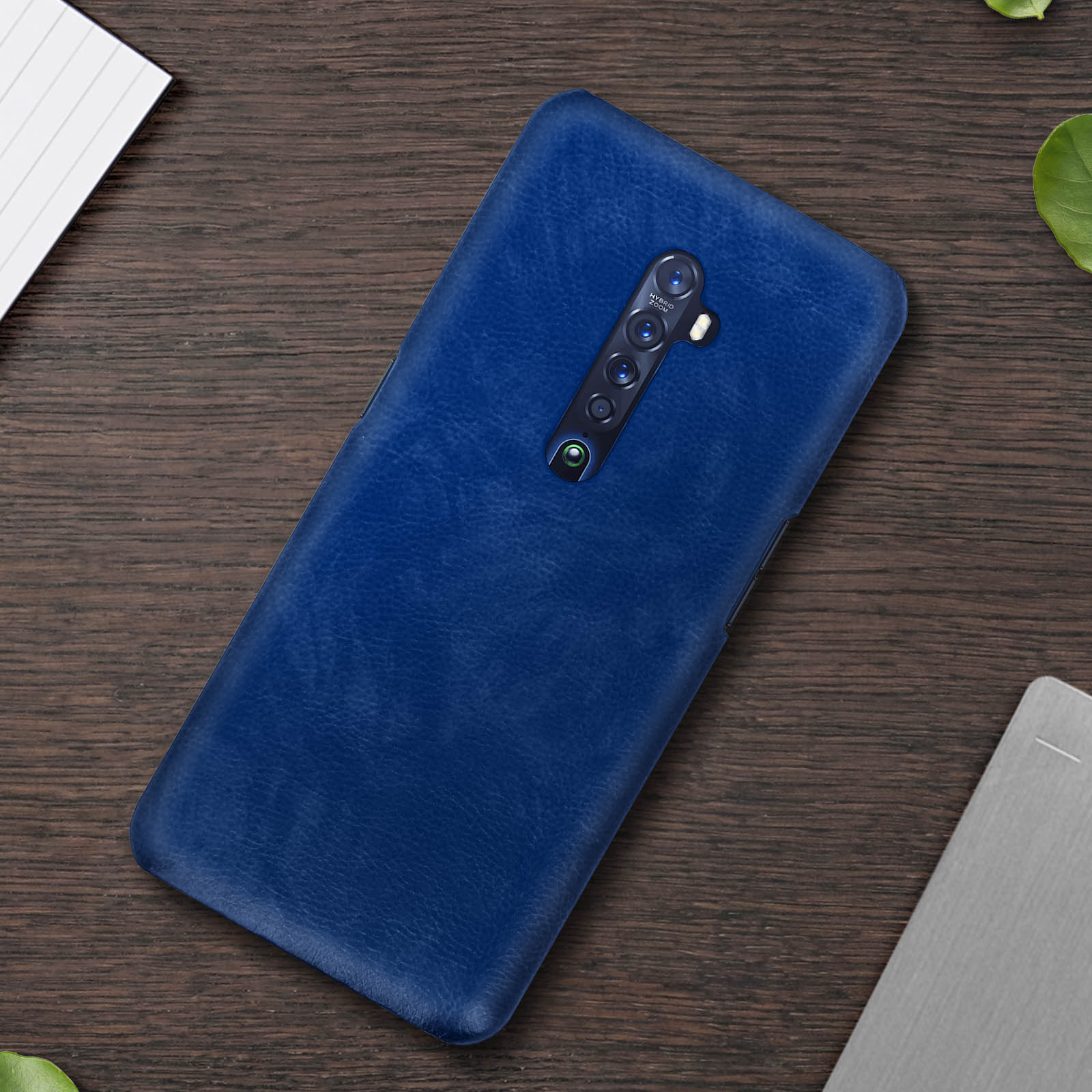 Custodia blu per telefono su un tavolo di legno. Sono presenti anche carta bianca e foglie verdi.