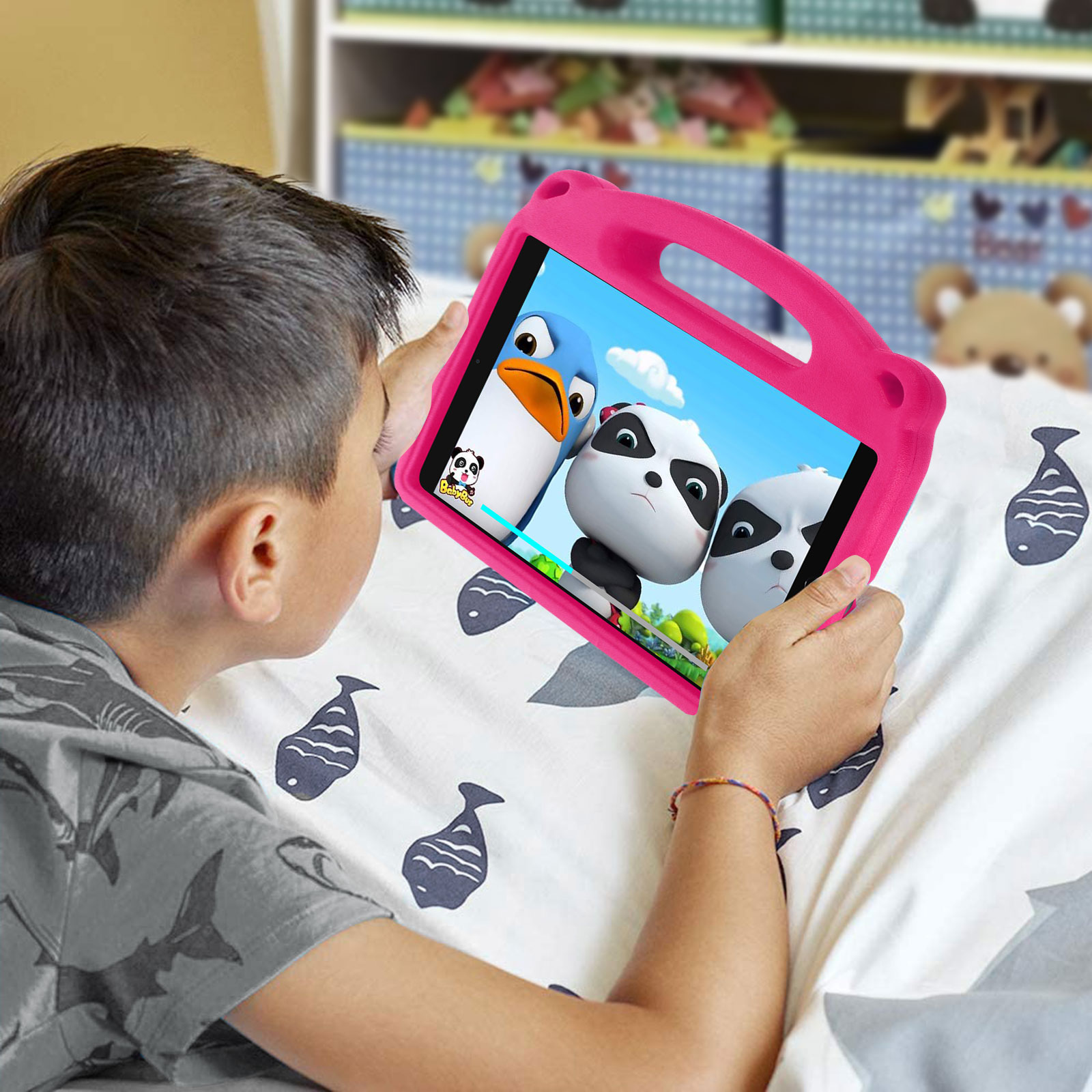 Kind schaut Zeichentrickfilm auf pinkem Tablet. Bett mit Fischmuster im Hintergrund.