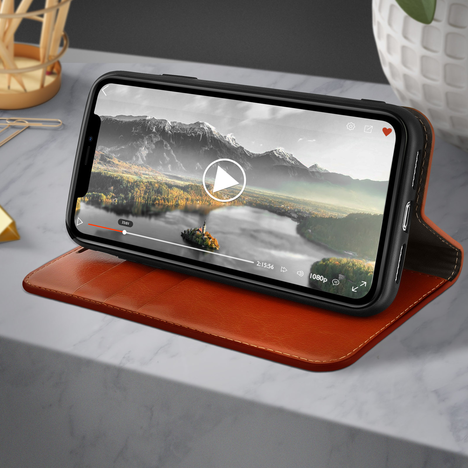 Een telefoon in een bruine hoes geeft een video van een landschap weer. De hoes staat op een marmeren oppervlak.