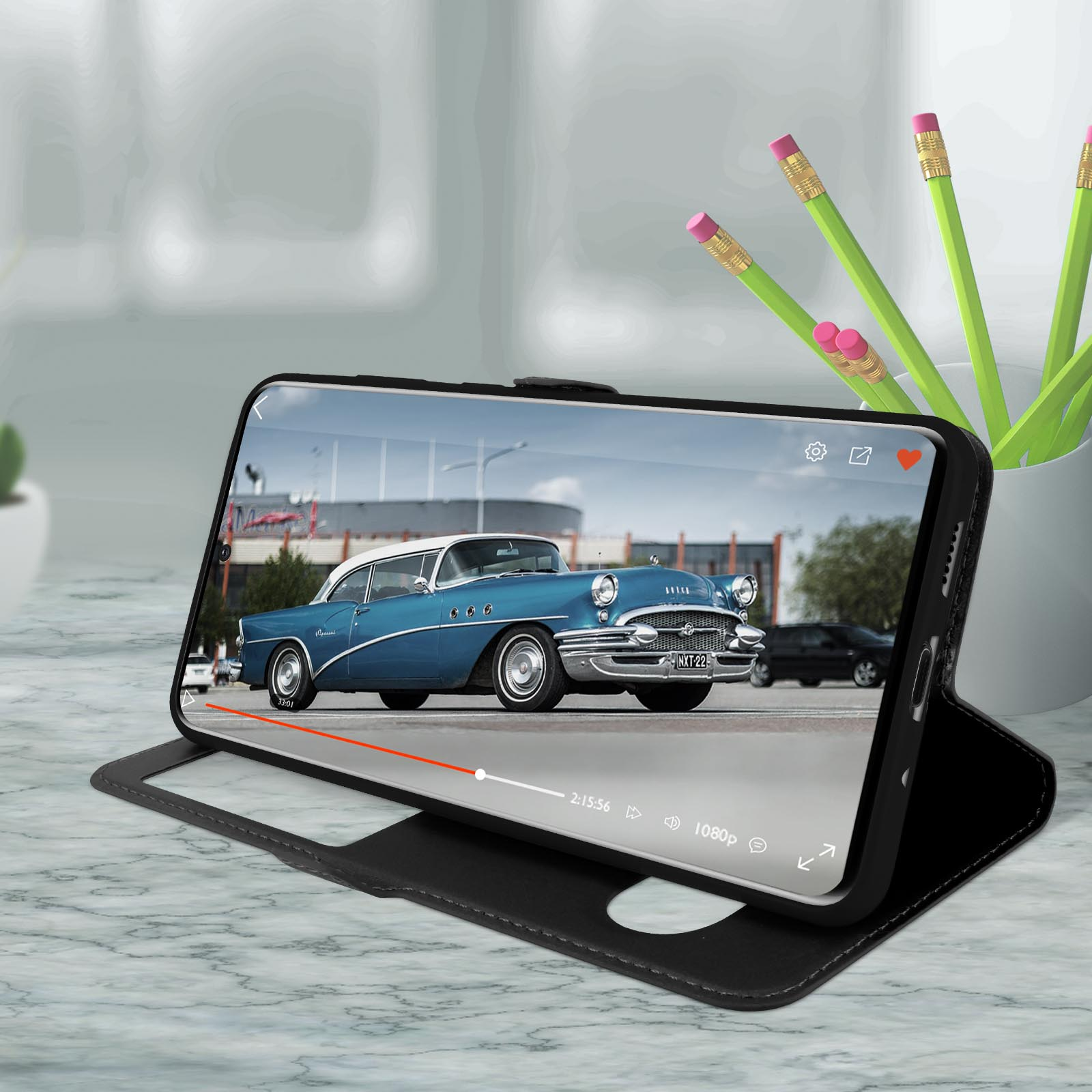 Smartphone in custodia nera, riproduce video su tavolo di marmo, matite sullo sfondo.