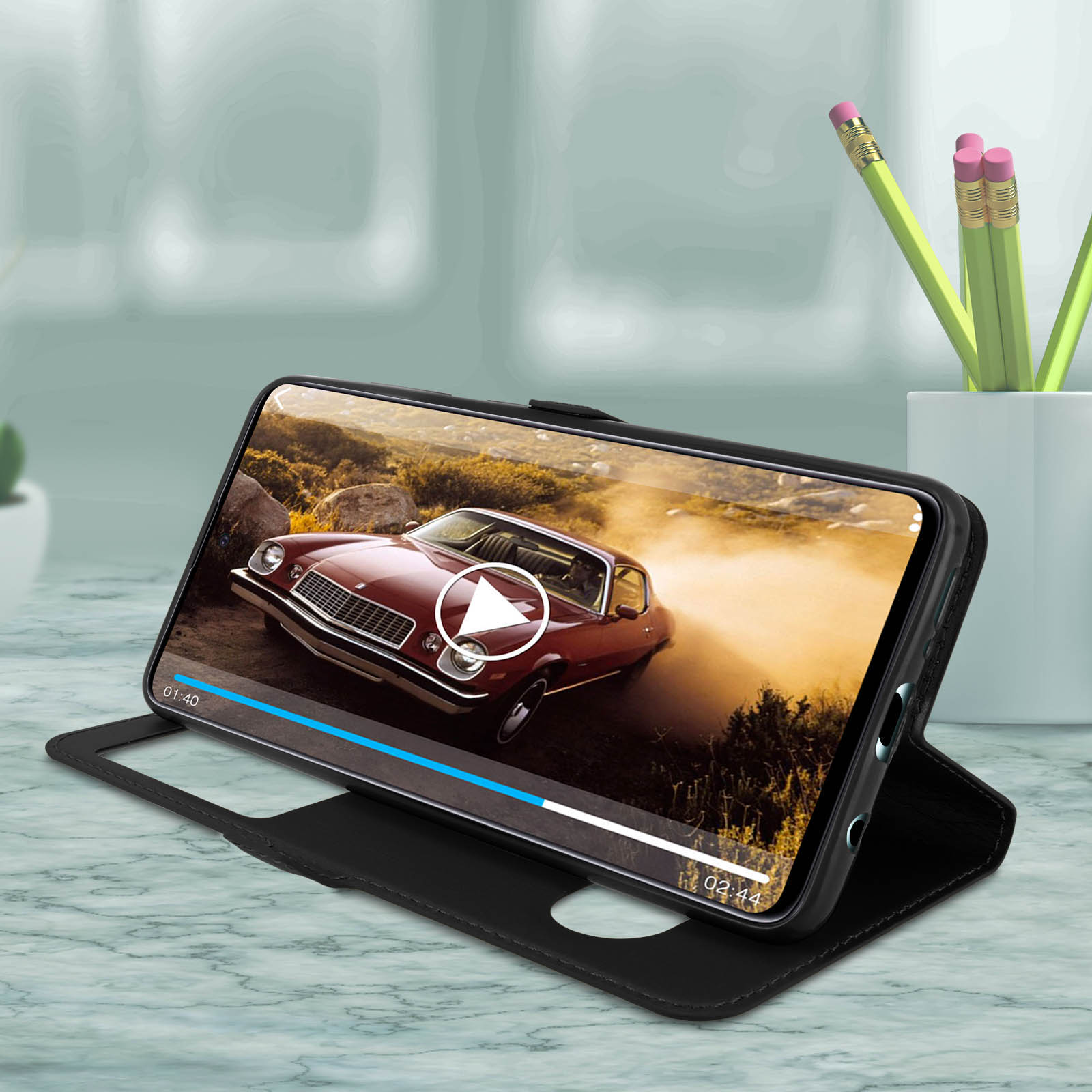 Un telefono con un video di un'auto è appoggiato in una custodia nera. Matite e un vaso sono sullo sfondo.