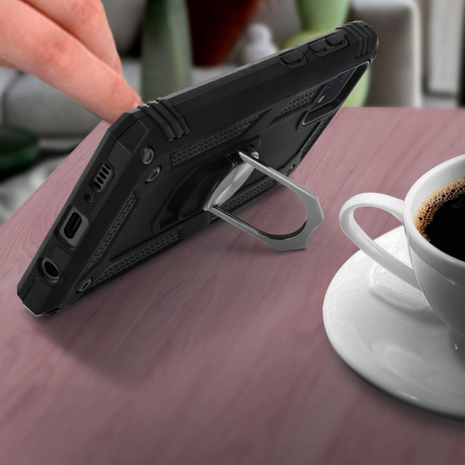Étui de téléphone noir avec support de bague, sur une table en bois. Tasse de café en arrière-plan.