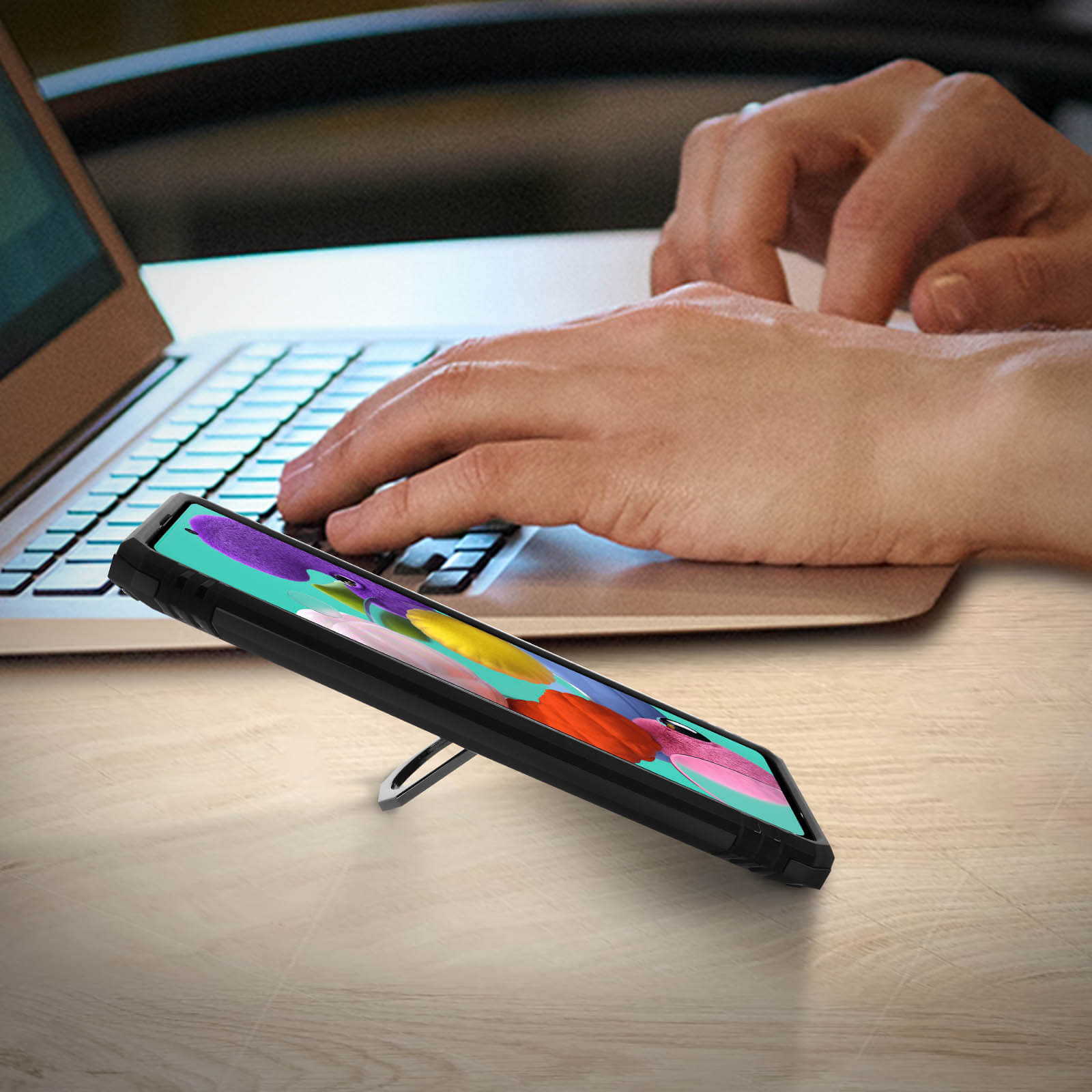 Un téléphone dans un étui est posé sur un support, avec des mains tapant sur un ordinateur portable en arrière-plan.