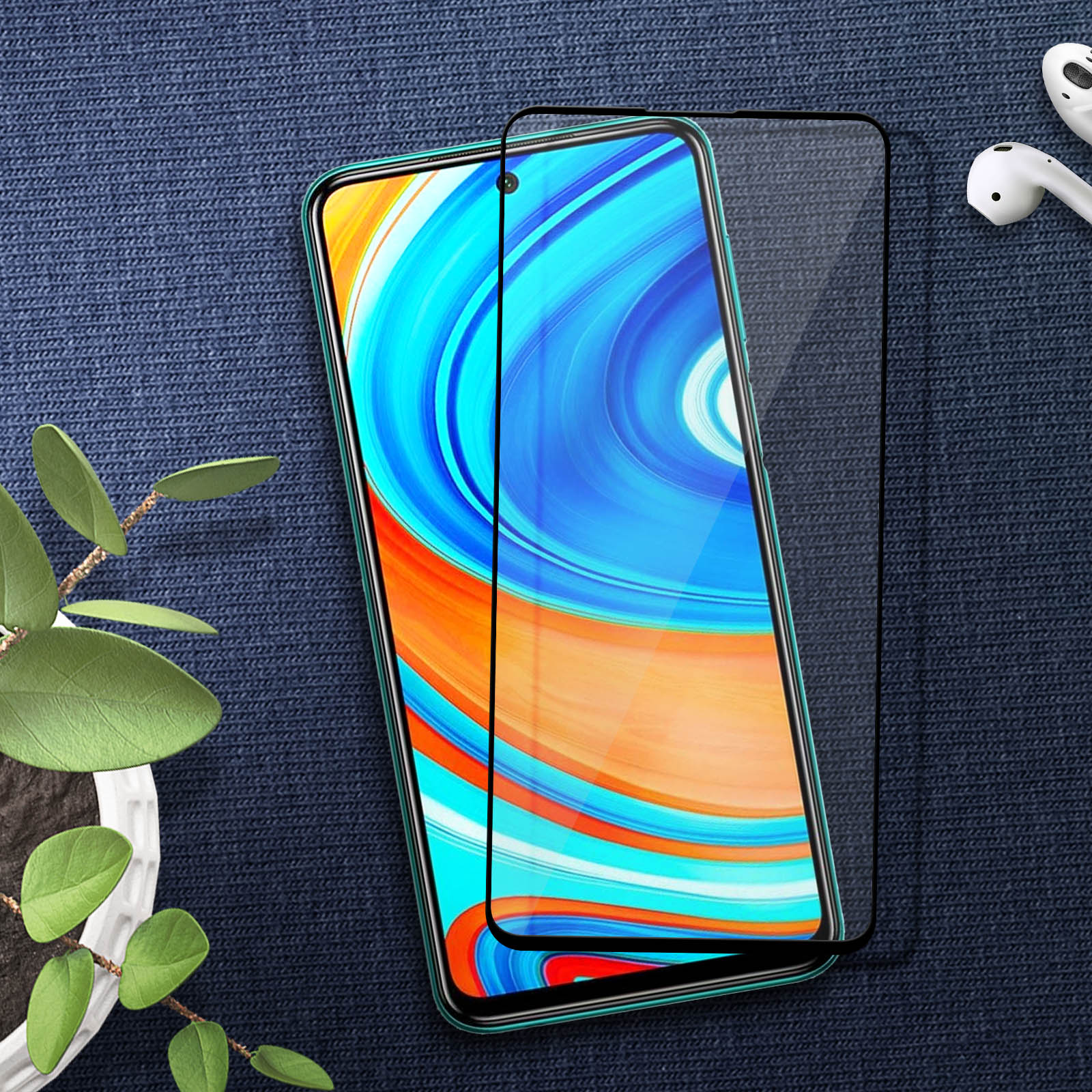 Smartphone con protezione schermo su tessuto blu. Una piccola pianta e auricolari wireless sono nel fotogramma.