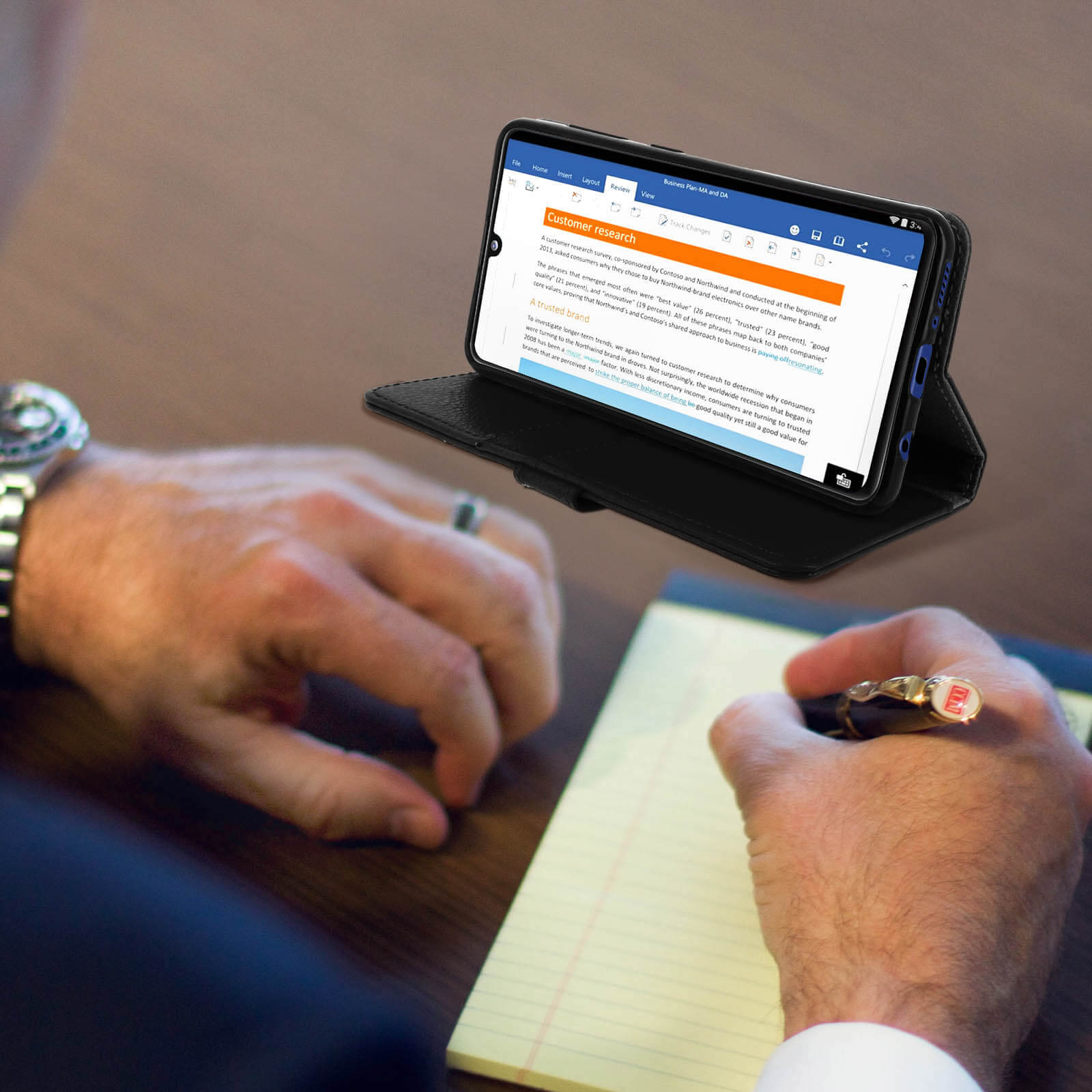 Une personne écrit sur un bloc-notes, avec un téléphone affichant un document en arrière-plan.