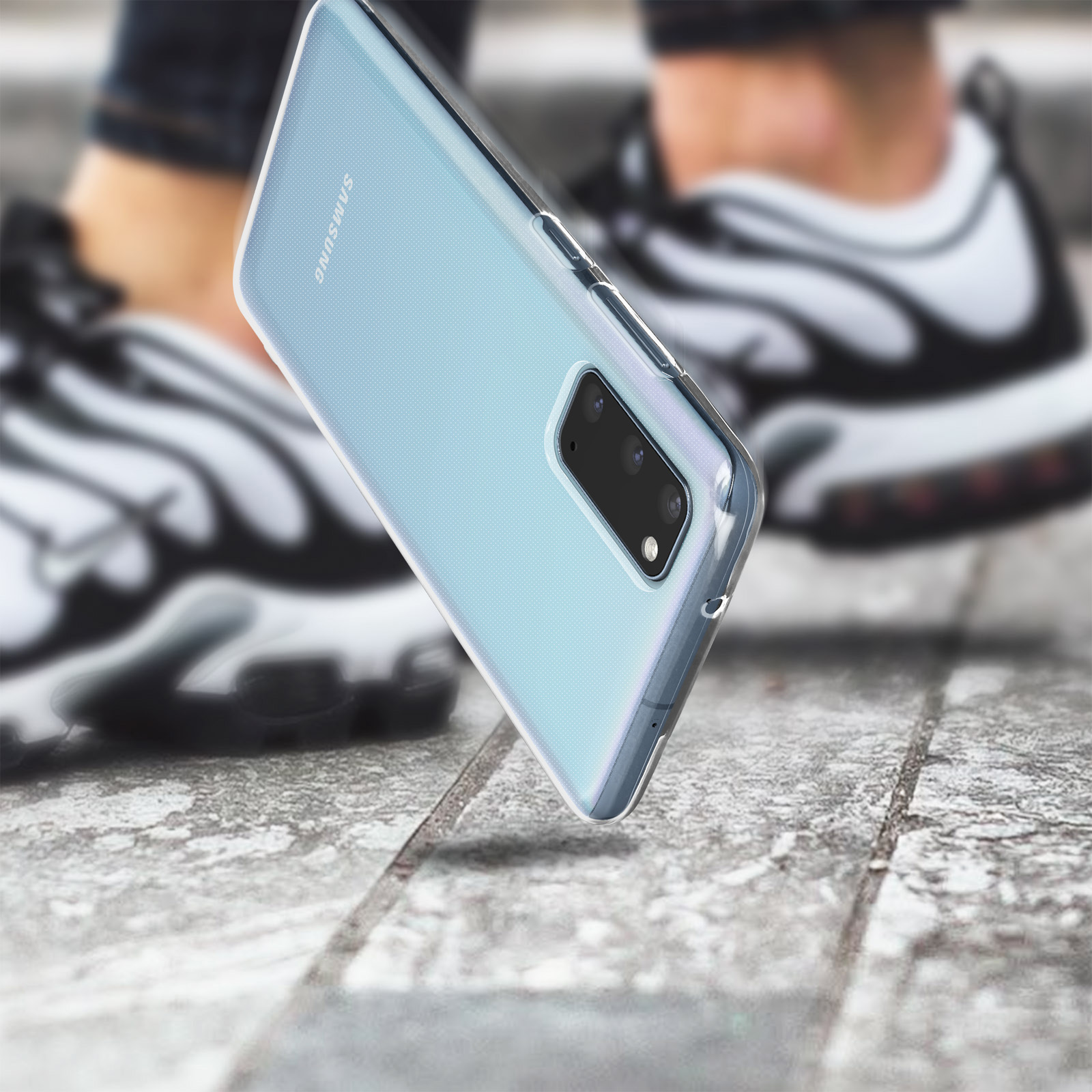 Un téléphone dans une coque transparente, tenu devant les jambes d'une personne. Fond : trottoir.