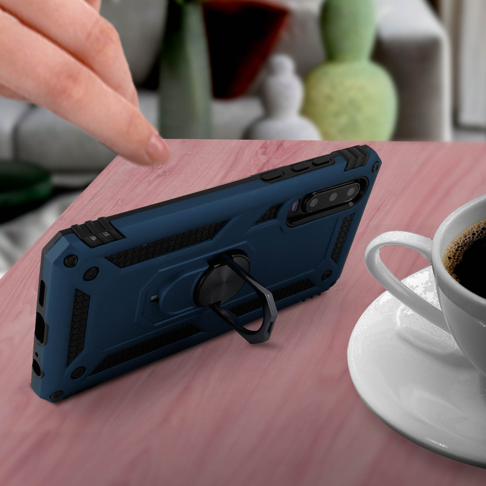 Een hand die een blauwe telefoonhoes vasthoudt met een ringhouder op een roze oppervlak, met een kopje koffie.