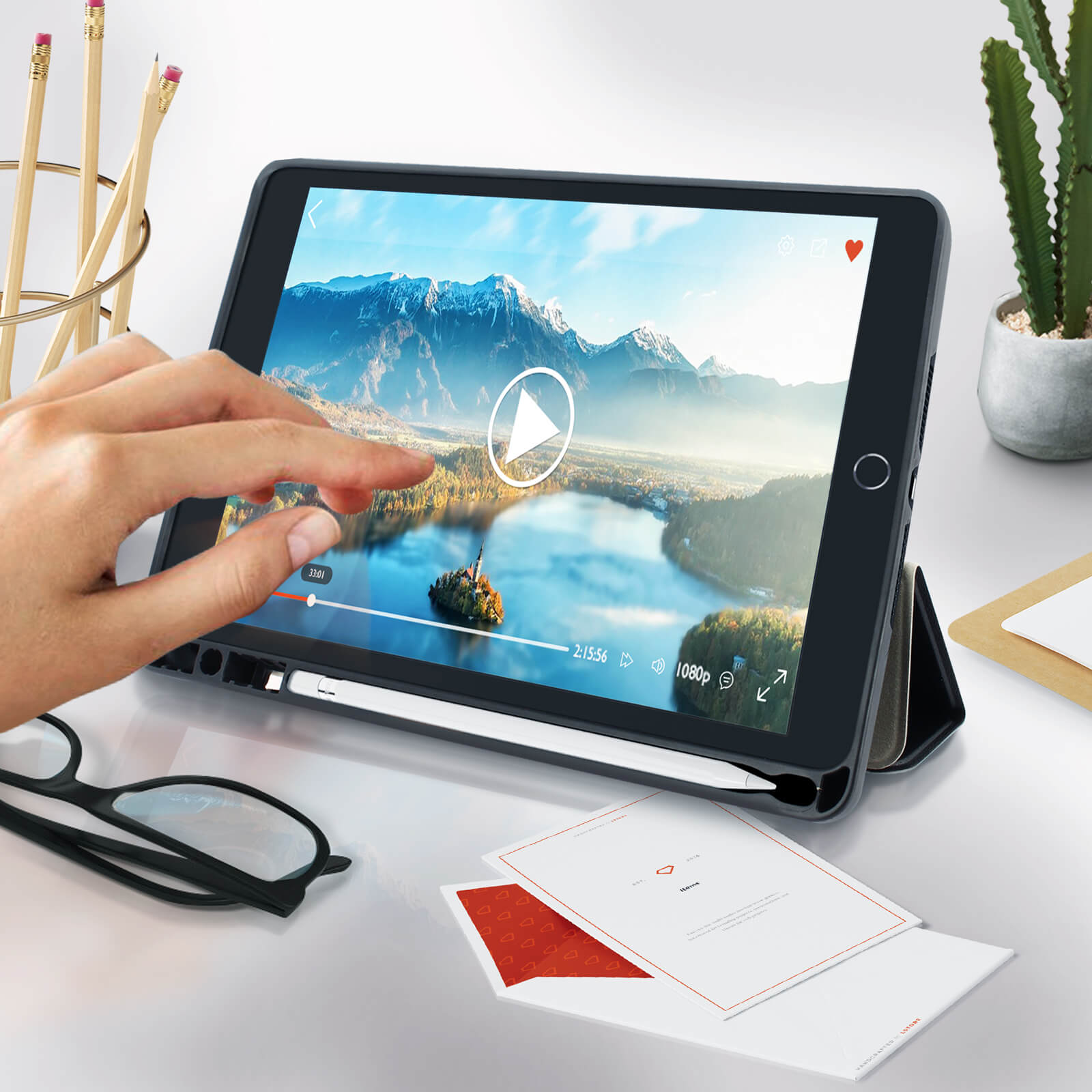 Tablet con video in riproduzione, mano che tocca lo schermo, matite, occhiali e fogli su un tavolo bianco.