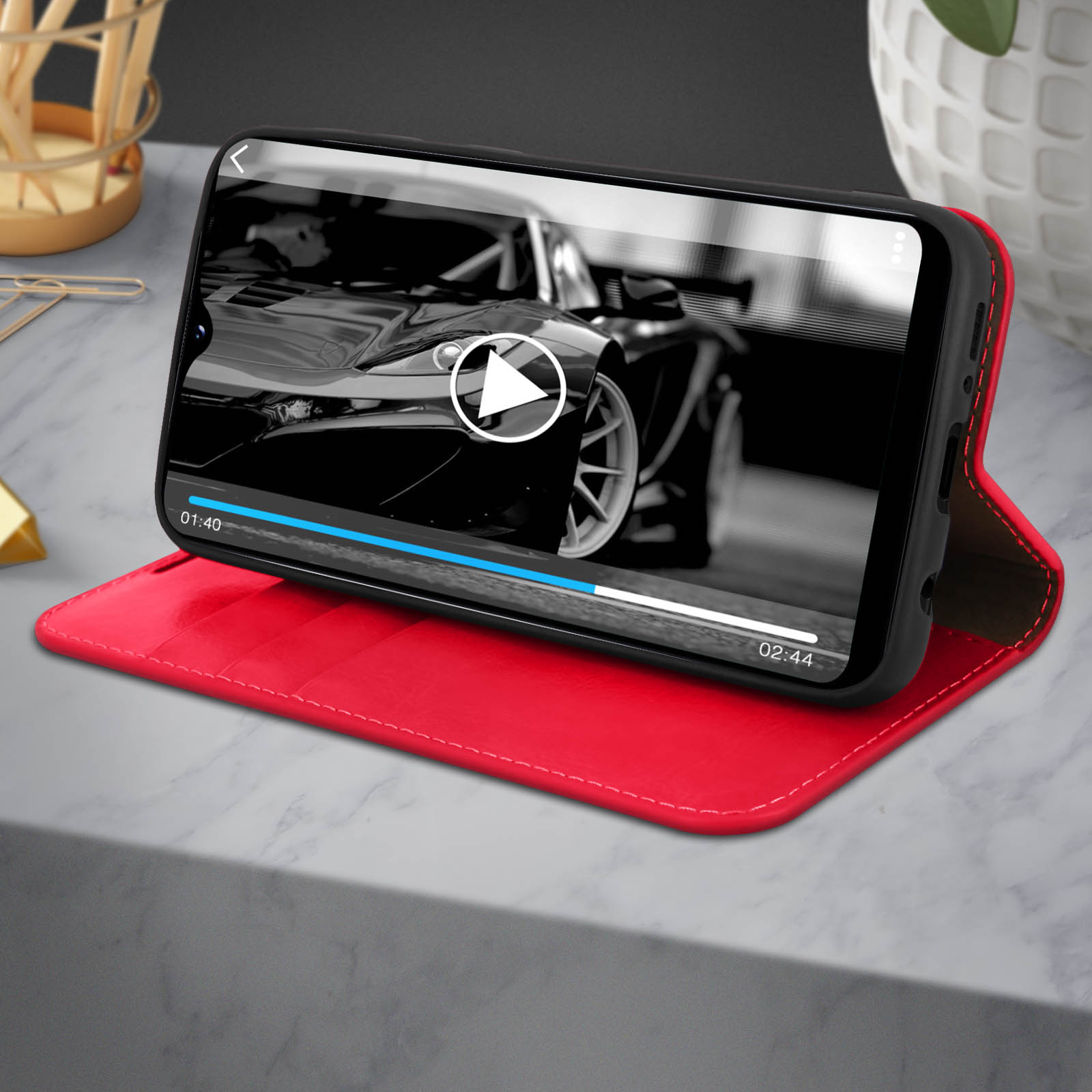 Telefon in roter Hülle, das ein schwarz-weißes Autovideo anzeigt. Die Hülle liegt auf Marmor.