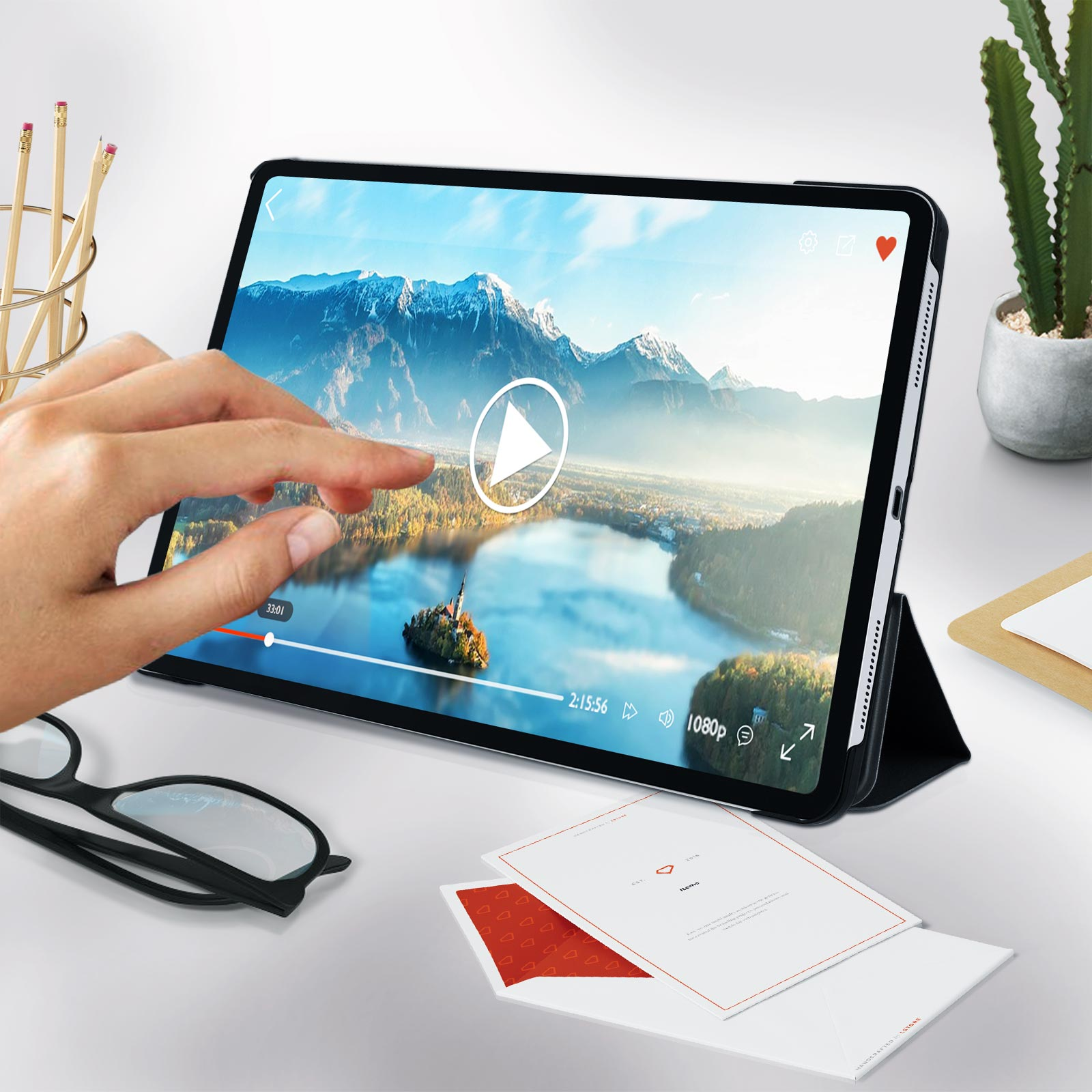 Tablet mostra un video, con pulsante play. Mano tocca lo schermo. Fogli e matite sul tavolo.