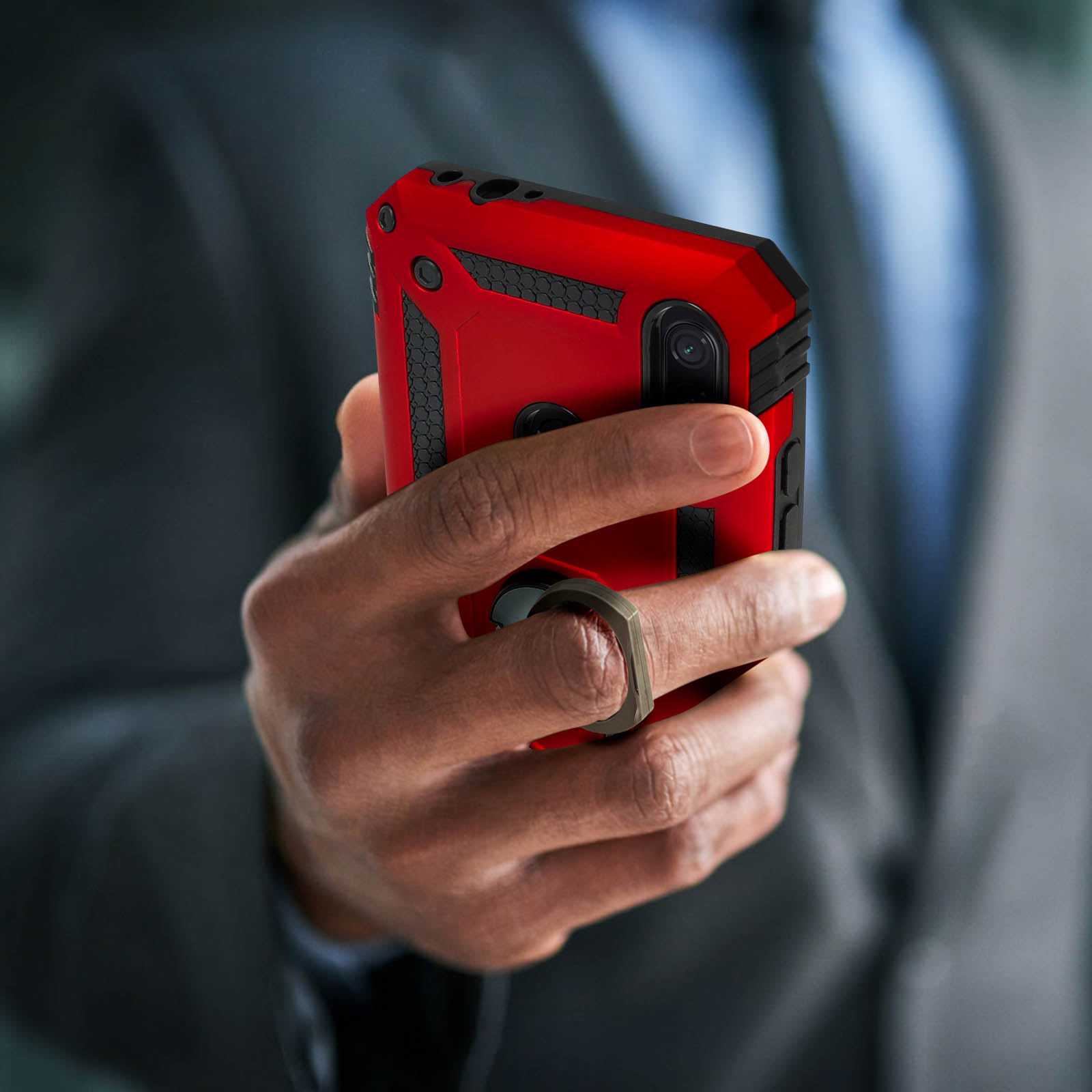 Une main tenant une coque de téléphone rouge avec un support de bague, avec un costume en arrière-plan.