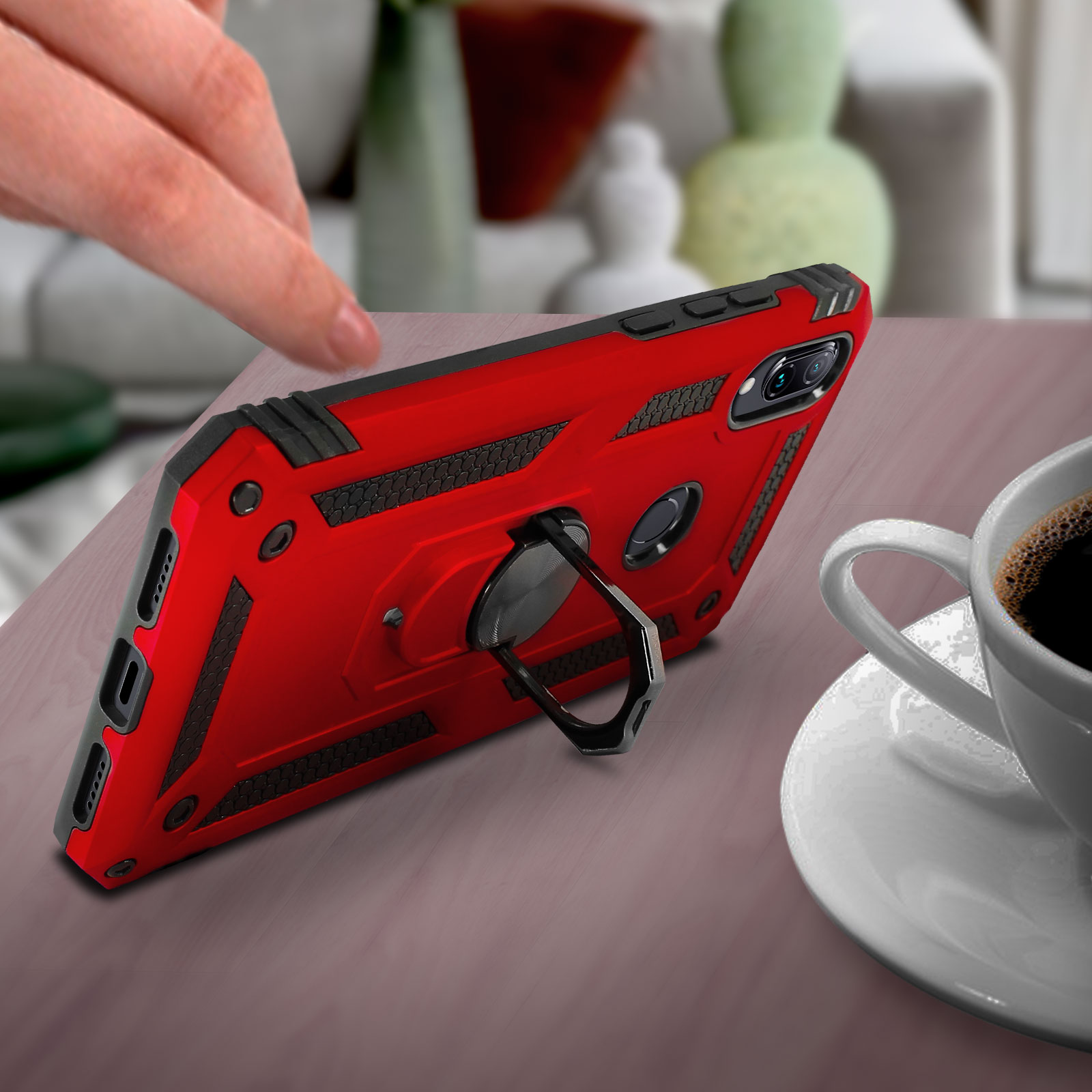 Une coque de téléphone rouge avec un support en anneau noir repose sur une table, avec une tasse de café et une main.