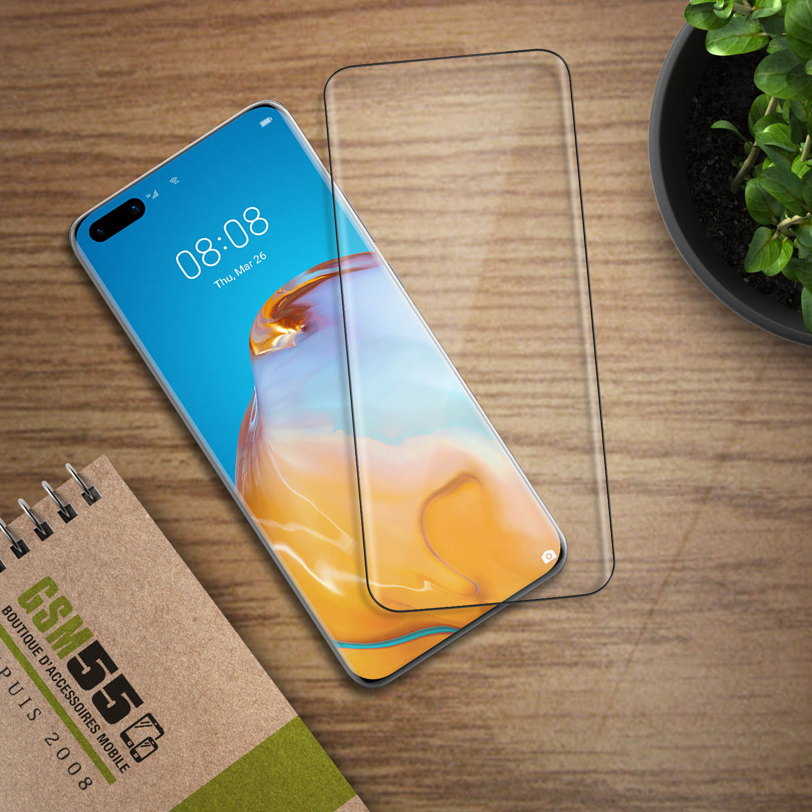 Uno smartphone con una protezione schermo è su un tavolo di legno. Un blocco note e una pianta sono presenti.