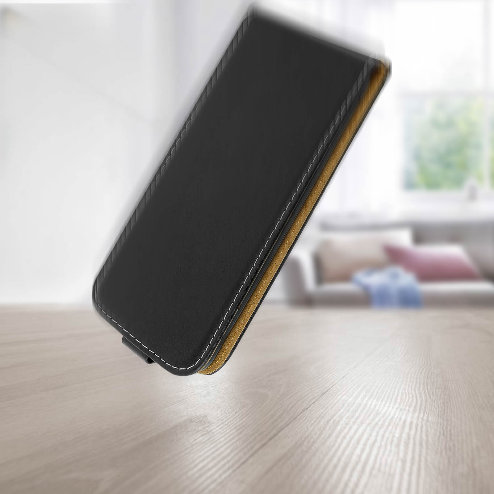 Una custodia nera per telefono cade su una superficie di legno. Sfondo: divano e finestra.