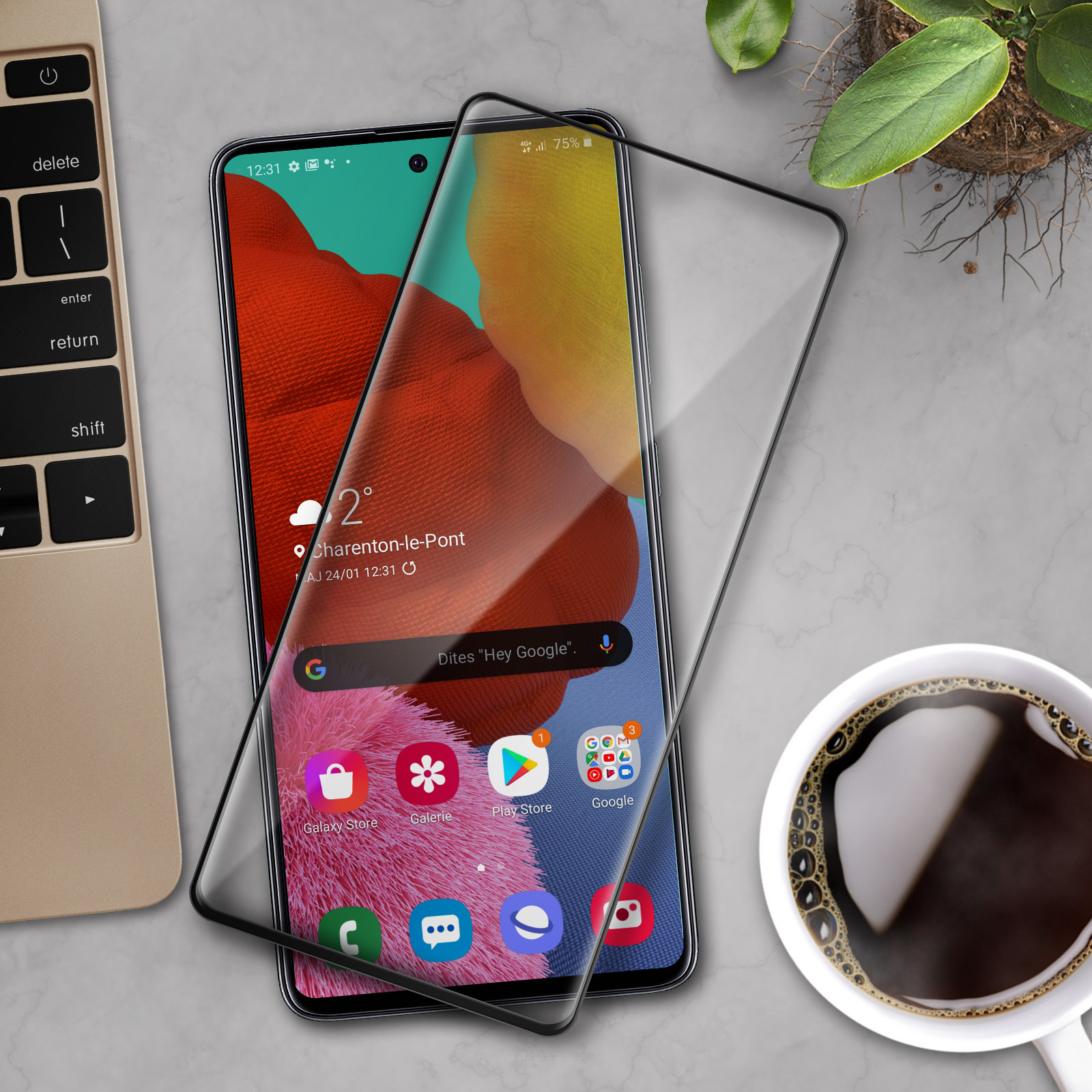 Uno smartphone con protezione schermo, che mostra app. Accanto, una tastiera per laptop e una tazza di caffè. Lo sfondo è una superficie in marmo.