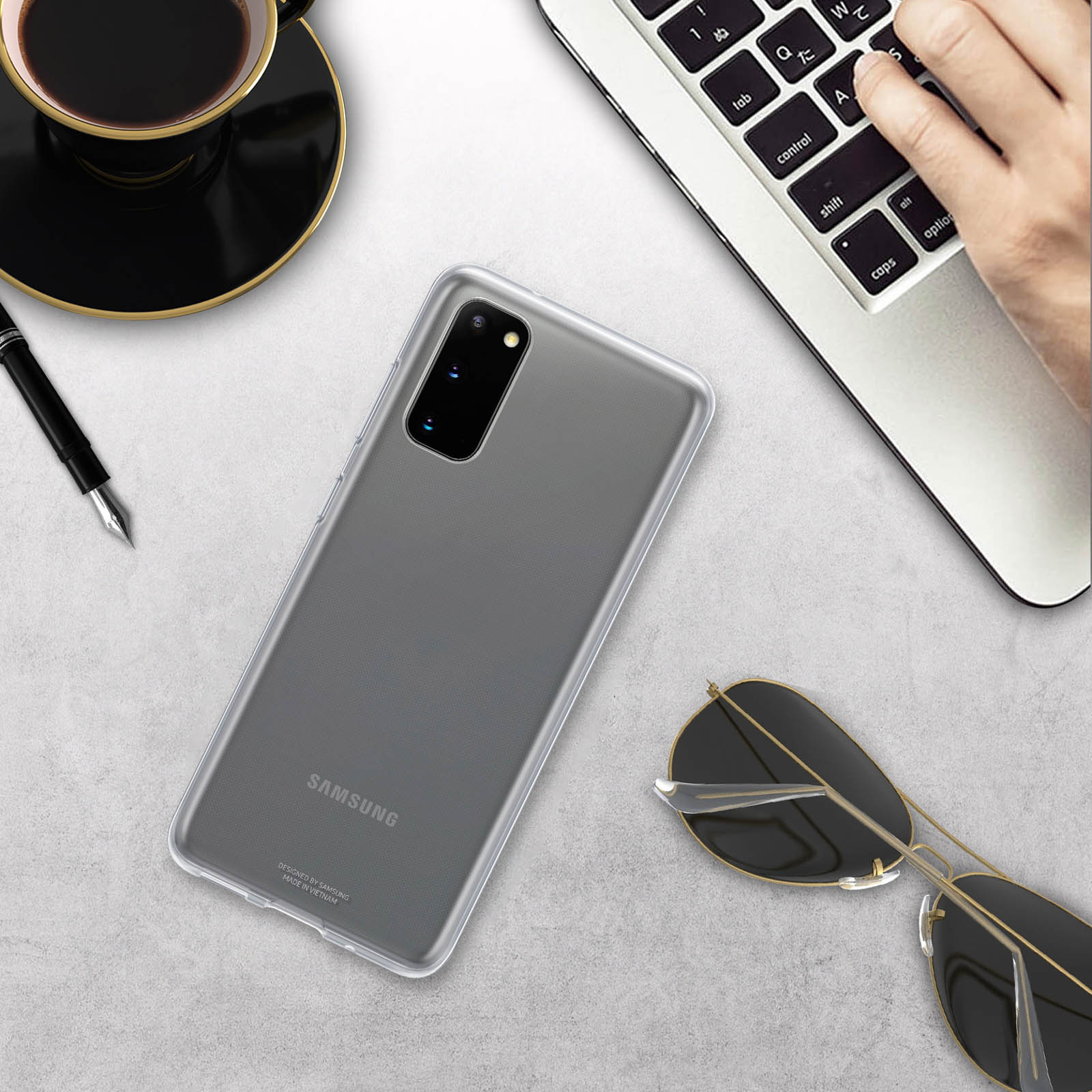 Una custodia trasparente per telefono, occhiali da sole, una penna e una tazza di caffè su una superficie grigio chiaro.