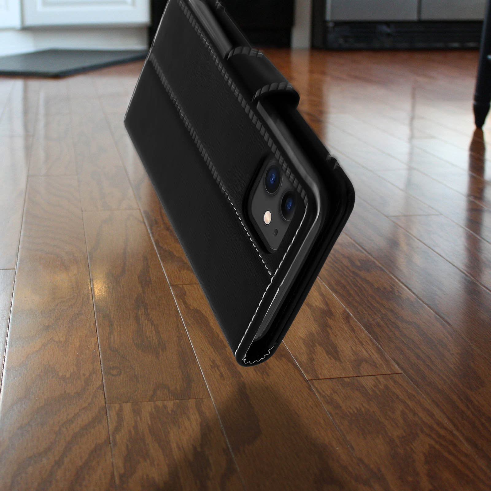 Custodia per telefono nera, chiusa, appoggiata su pavimento in legno. Dettagli di cucitura bianchi. Mostra fotocamera e flash.