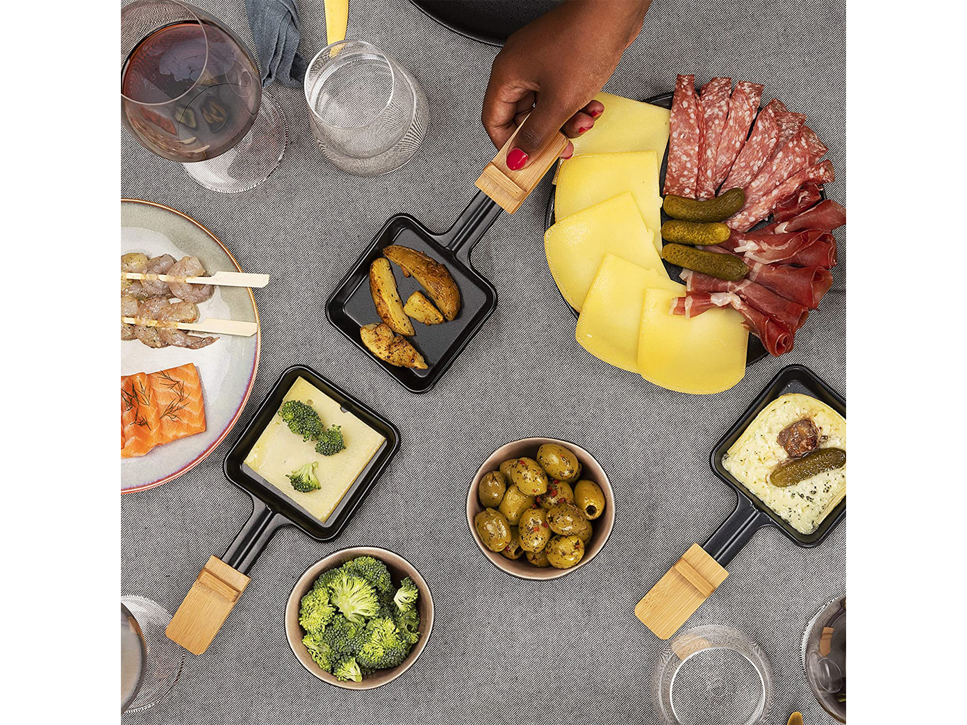 Eine Draufsicht auf einen Tisch, der für eine Mahlzeit mit Raclette-Pfannen, Essen und Getränken gedeckt ist.