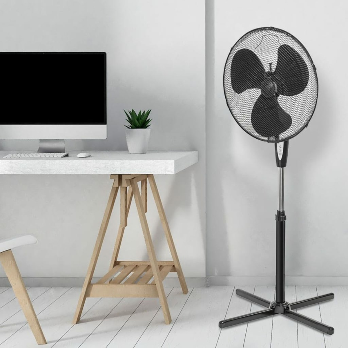 Czarny wentylator na stojaku, w białym pokoju. Komputer i roślina na białym biurku.