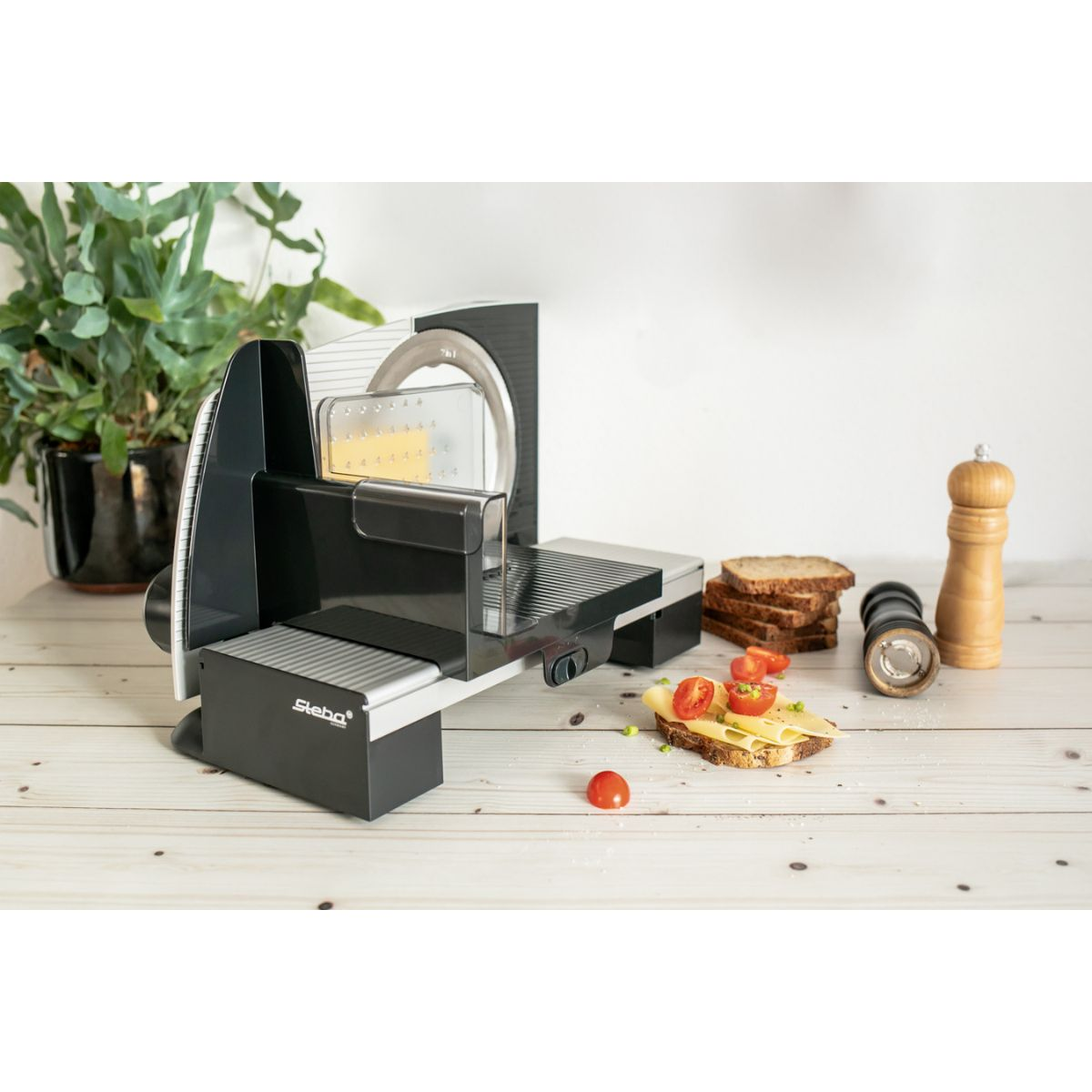 Eine schwarz-silberne Lebensmittelschneidemaschine mit Brot und Tomaten. Eine Pfeffermühle rechts.