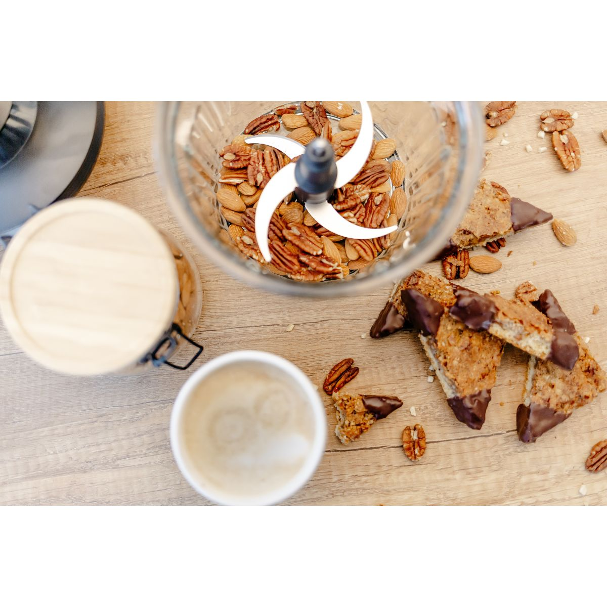 Ein Mixer mit Mandeln, Pekannüssen und Keksen steht auf einem Holztisch. Eine Tasse und ein Glas sind ebenfalls vorhanden.