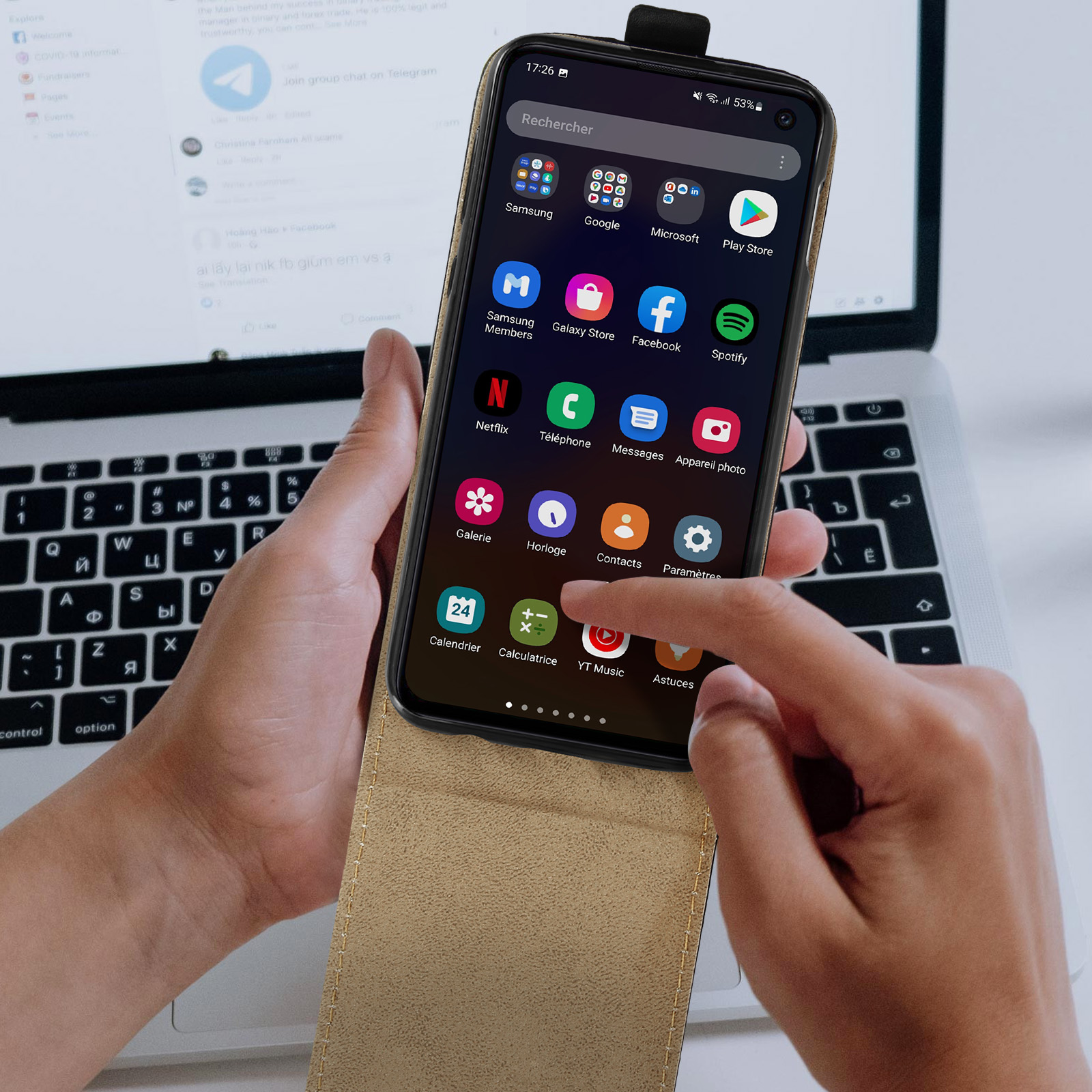 Una mano che tiene un telefono che mostra app sullo schermo, un laptop è aperto sullo sfondo.