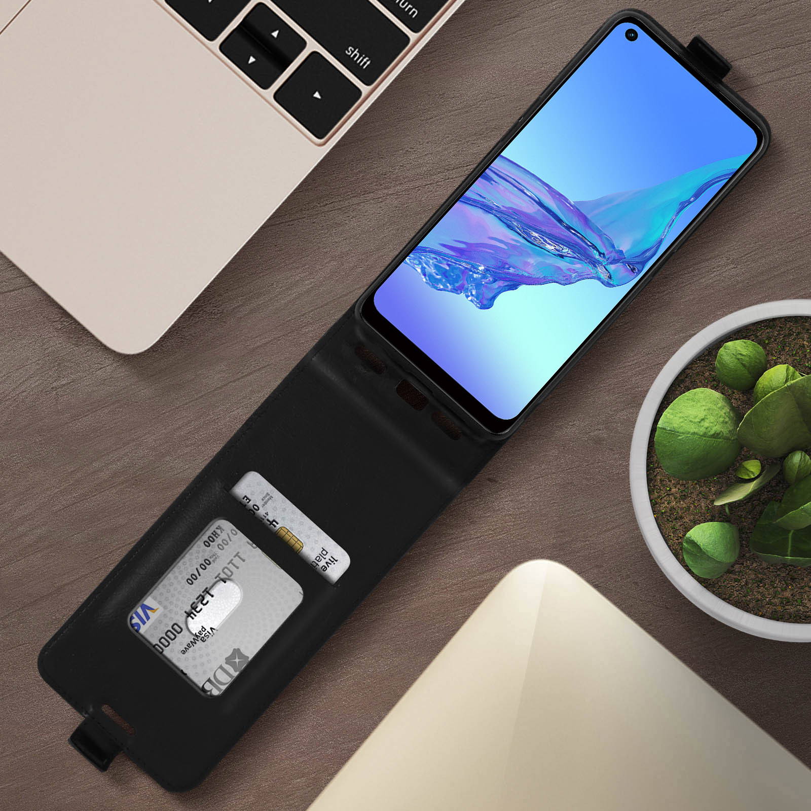 Un telefono in una custodia nera su una scrivania di legno, con un laptop, una pianta e una carta di credito.