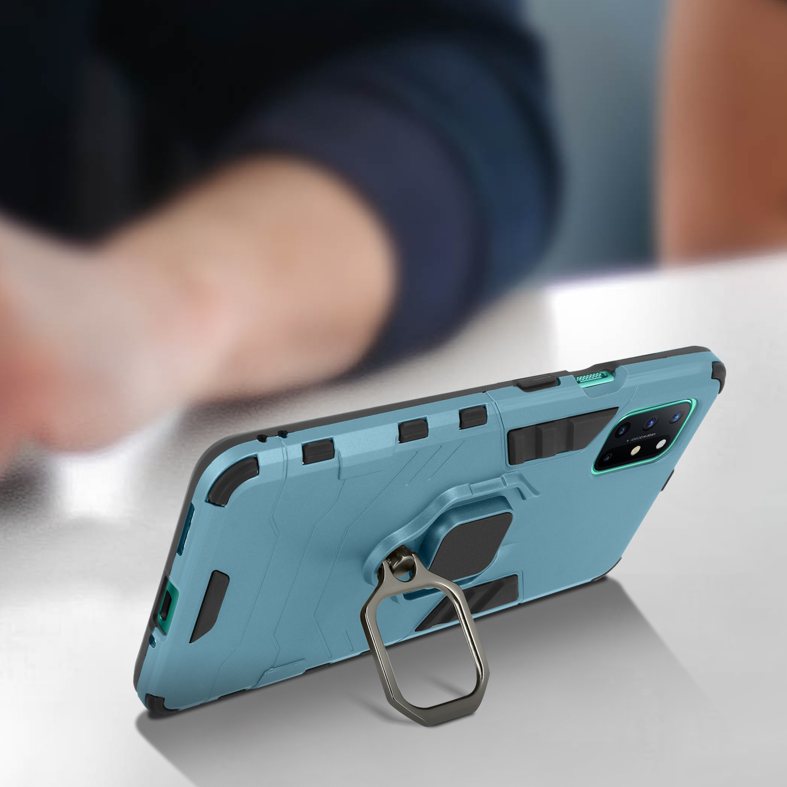 Custodia blu per telefono con anello in metallo. Una mano tiene il telefono. La custodia è su un tavolo bianco.