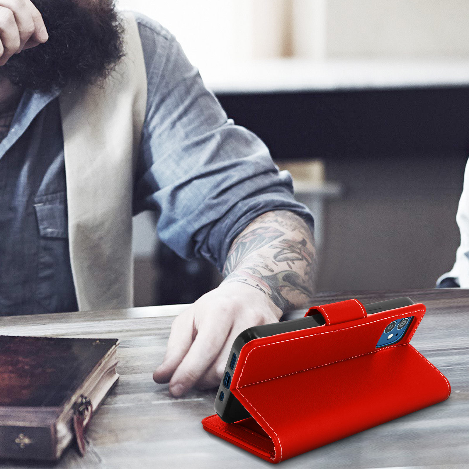 Un téléphone dans un étui rouge sur une table. Un homme barbu est à l'arrière-plan.