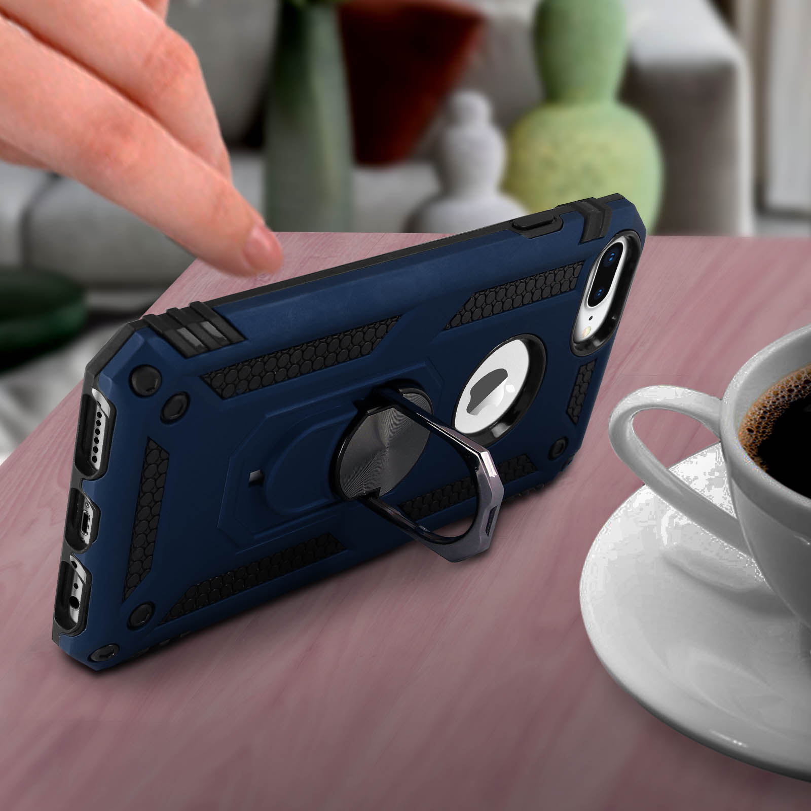 La main d'une personne près d'une coque de téléphone bleue avec un support annulaire sur une table. Une tasse de café à droite.