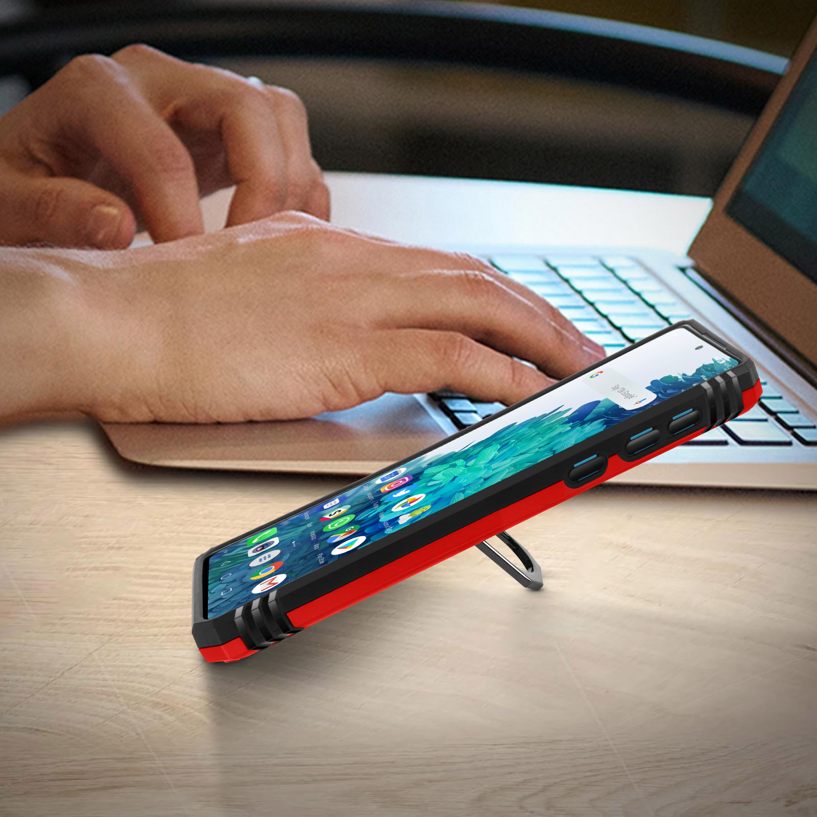 Telefoon in een rode en zwarte hoes, schuin op een bureau, met iemand die op een laptop typt.