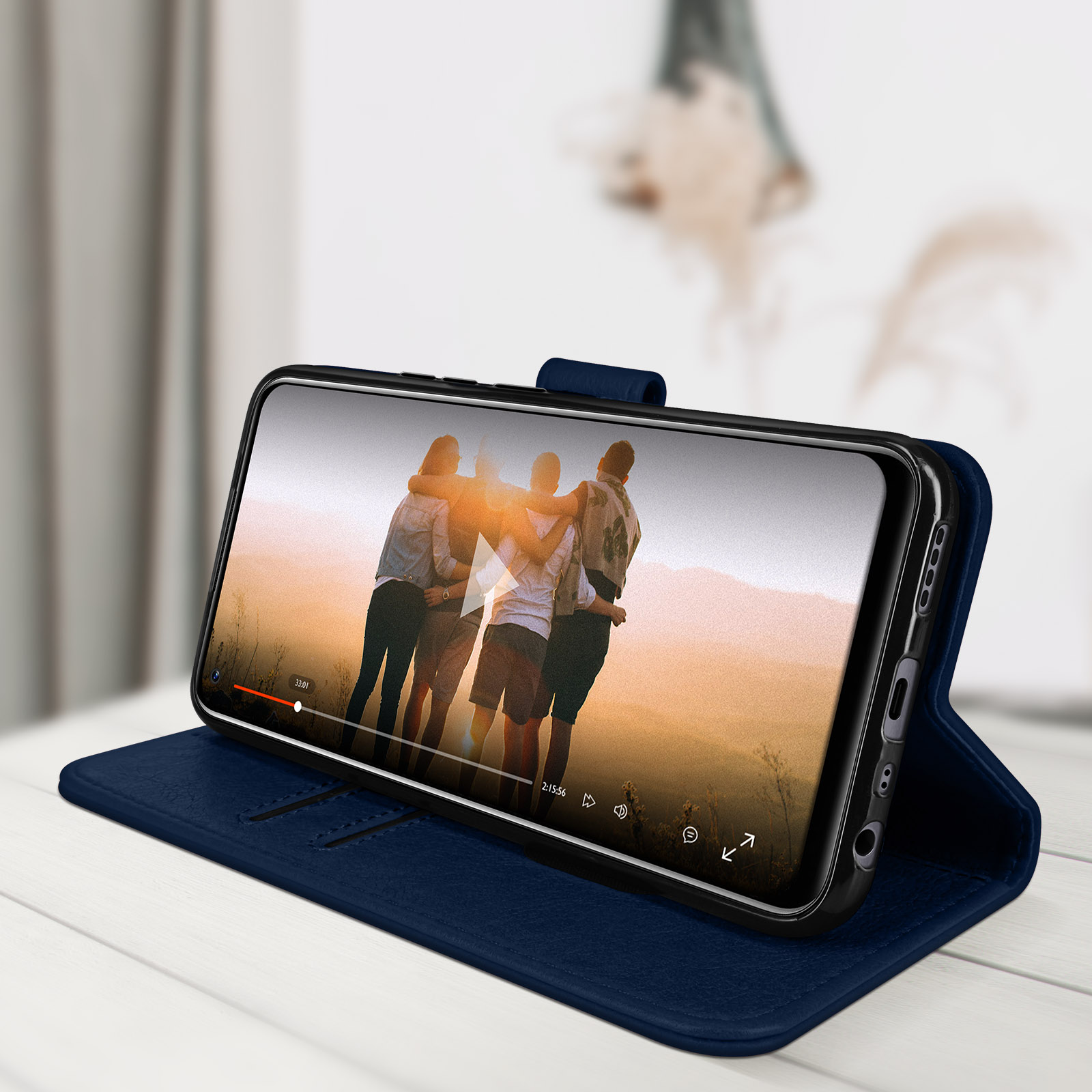 Telefon in blauem Etui, spielt Video. Das Etui ist offen. Personen sind auf dem Bildschirm. Das Etui liegt auf einem Tisch.