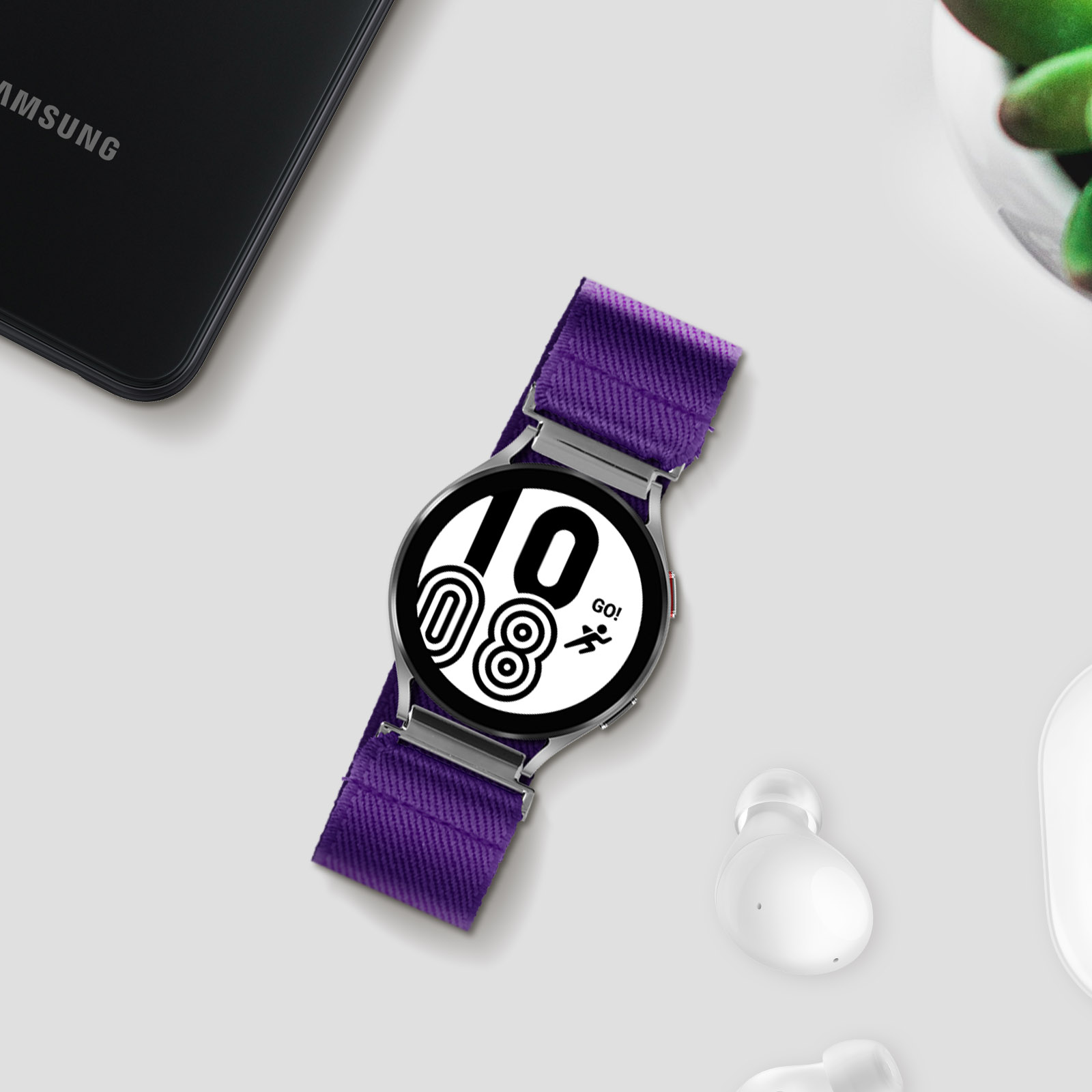 Un orologio Samsung Galaxy con cinturino viola, esposto su una superficie bianca con accessori.