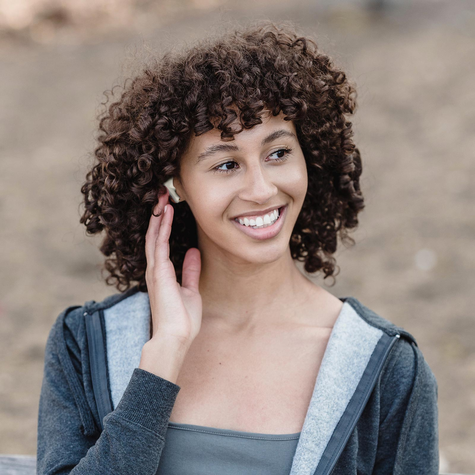 Una donna con i capelli ricci sorride, indossa auricolari. Lo sfondo è un parco sfocato.