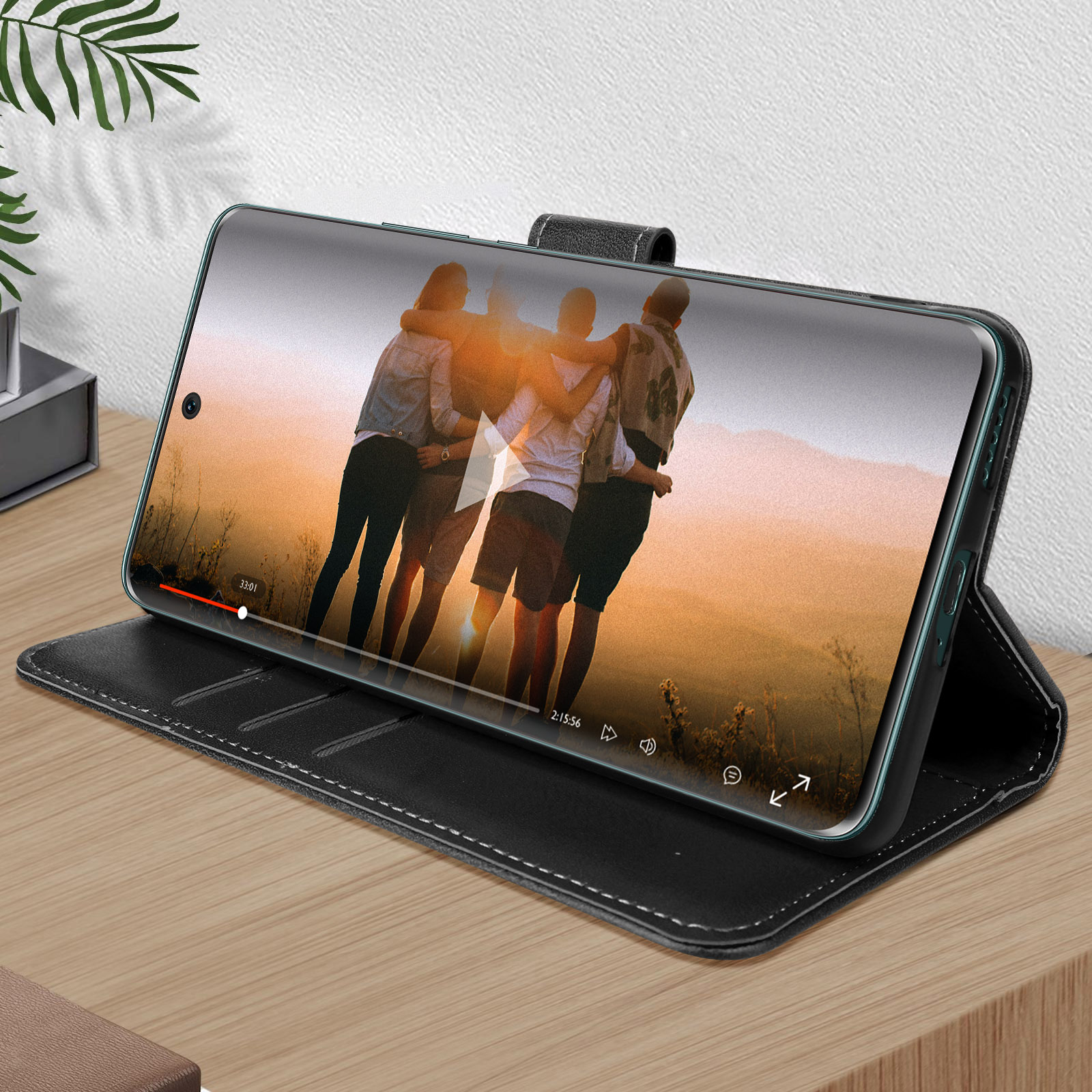 Telefono in custodia nera che riproduce video, su tavolo di legno. Persone sullo schermo.