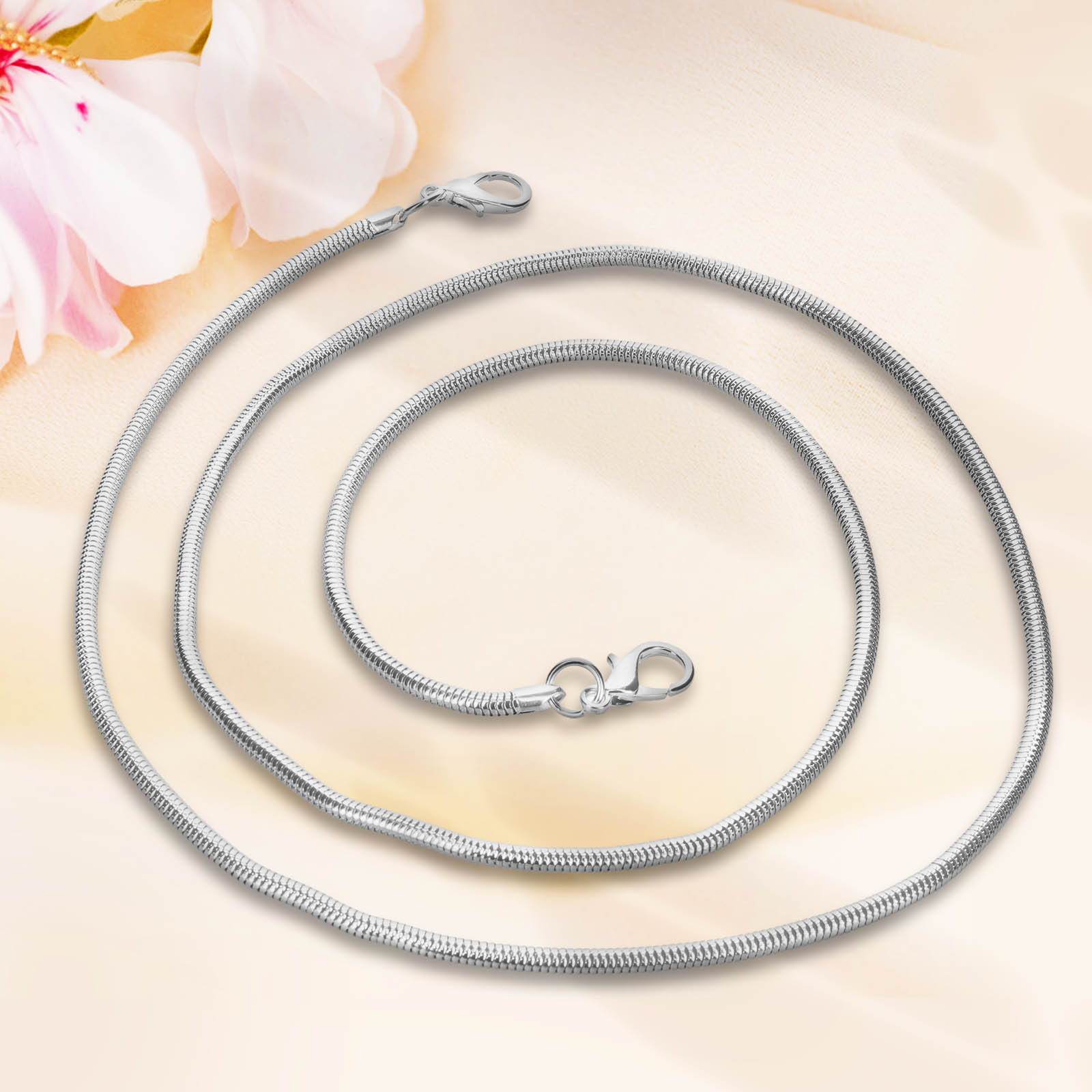 Una collana a catena di serpente argentata con chiusura a moschettone su sfondo crema con fiori rosa.