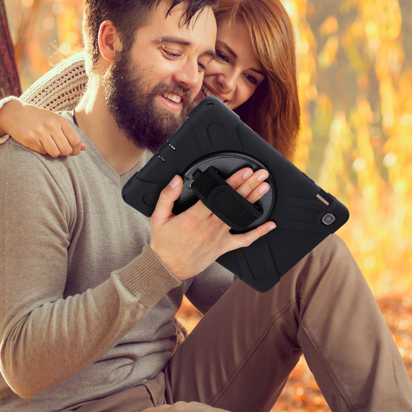 Ein lächelndes Paar hält ein schwarzes Tablet. Der Hintergrund sind verschwommene Herbstblätter.