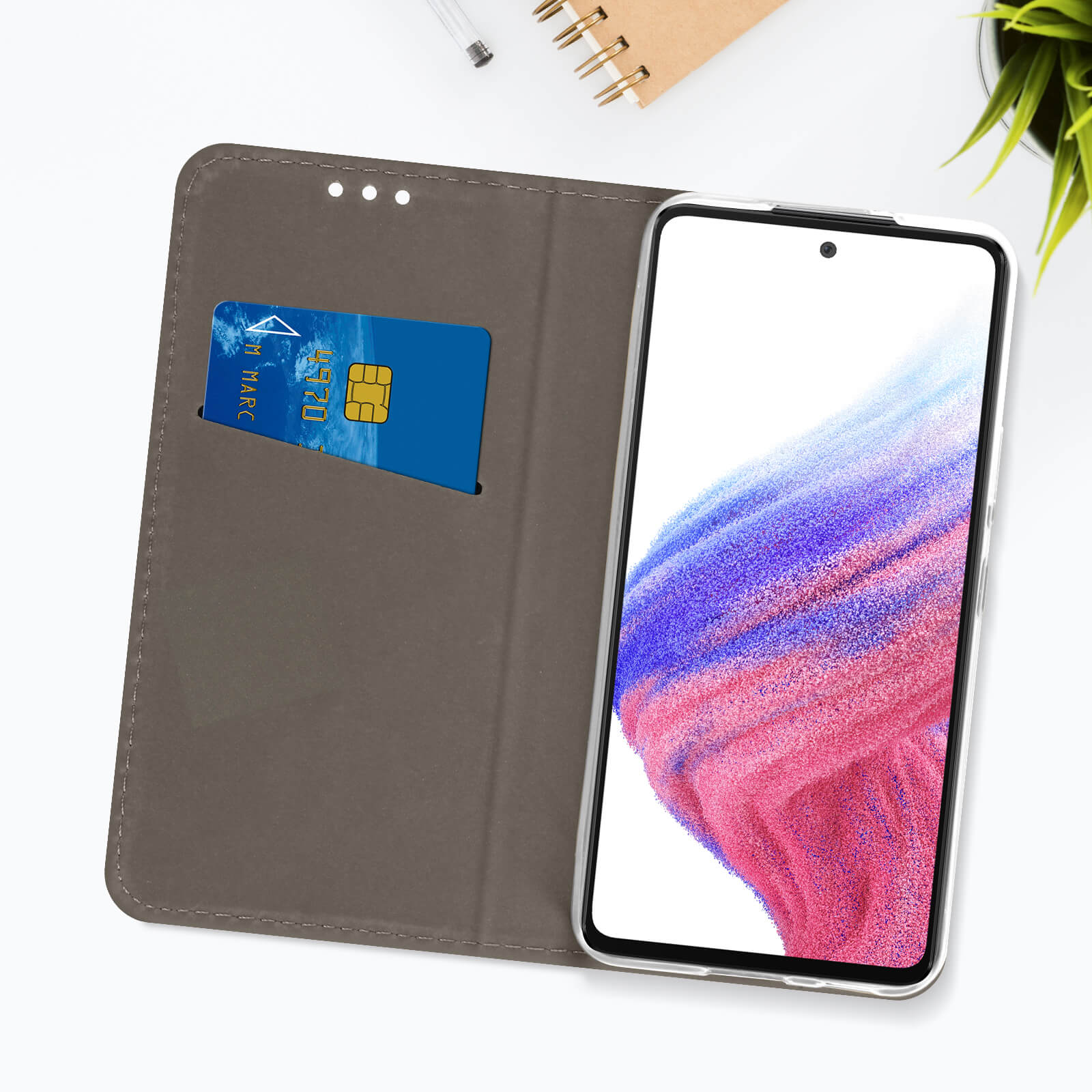 Téléphone dans un étui, portefeuille ouvert avec carte de crédit, sur surface blanche avec plante.